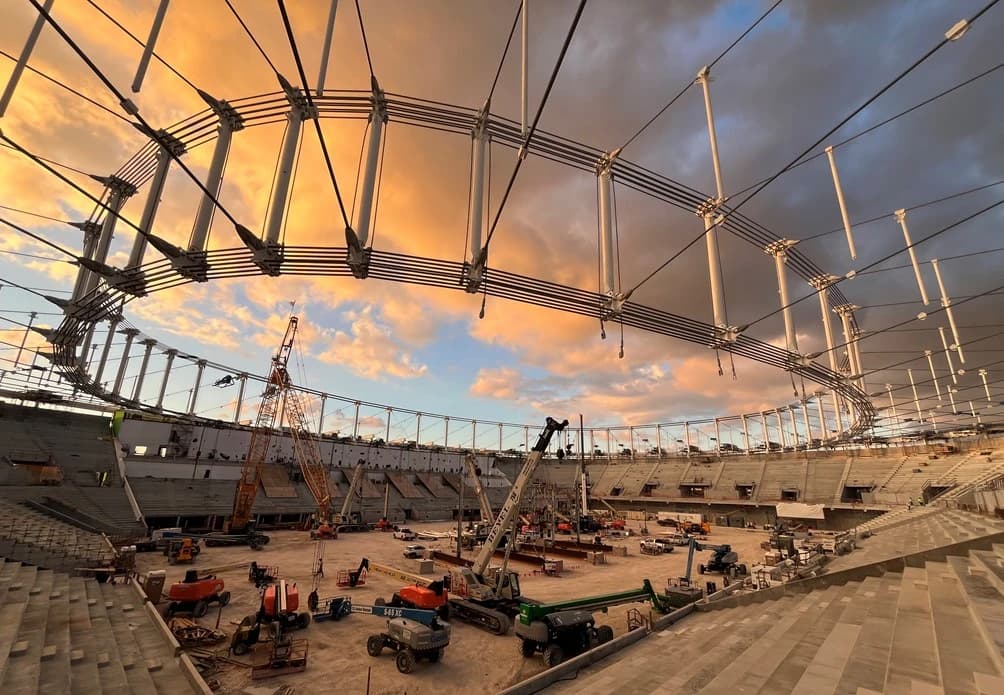 Un grand stade en construction au coucher du soleil, montrant des tribunes en béton, de multiples grues et engins de chantier au sol, et l'ossature métallique du toit sous un ciel aux teintes orange et grises.