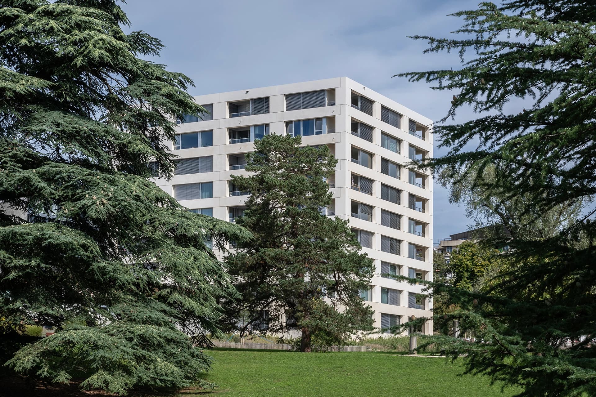 Un bâtiment moderne beige clair avec de nombreuses fenêtres et stores extérieurs est partiellement masqué par de grands arbres à feuilles persistantes vert foncé, au-dessus d'une pelouse verdoyante sous un ciel bleu clair.