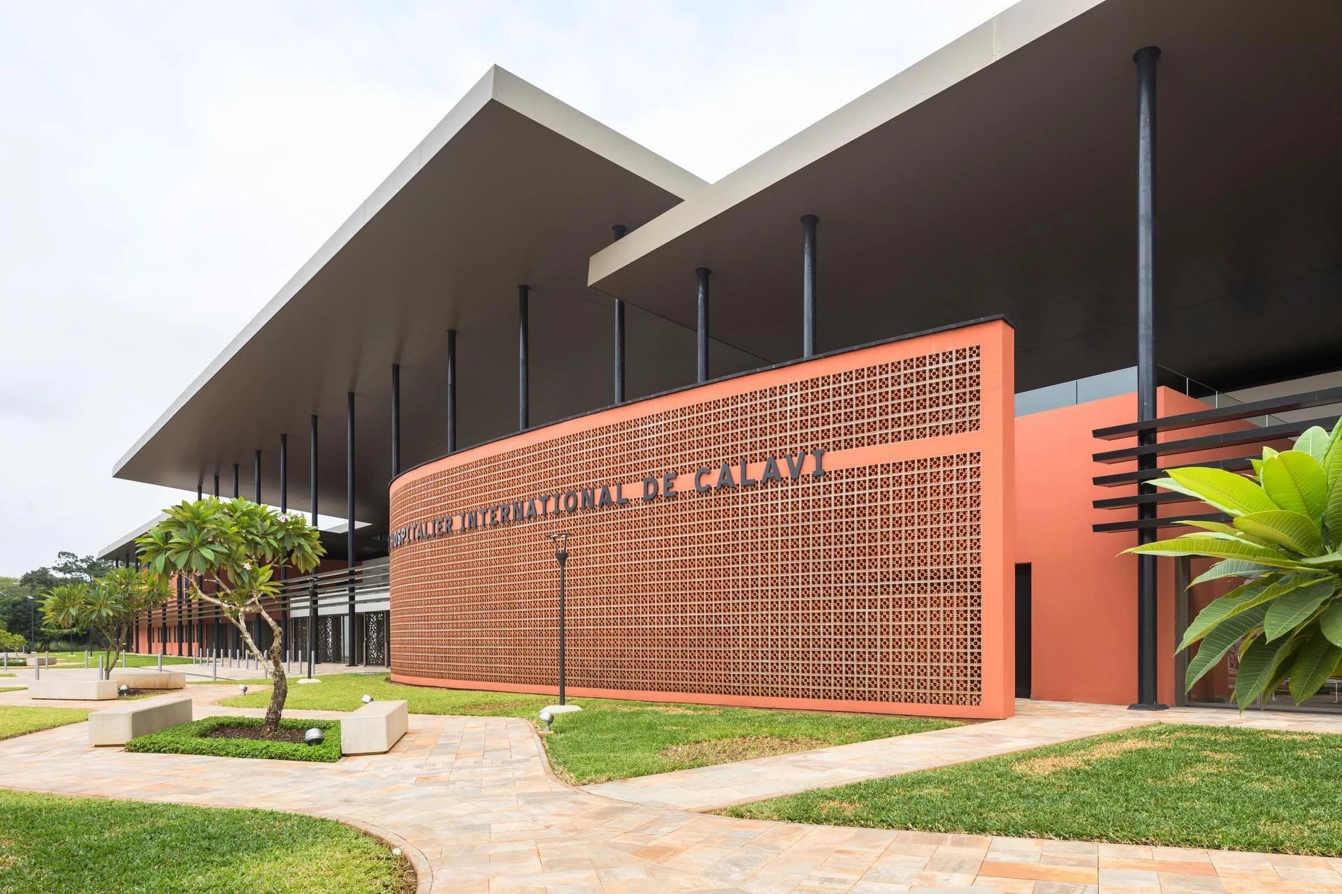 Façade incurvée d'un bâtiment moderne de couleur orange-terracotta avec un motif ajouré, portant l'inscription "HÔPITALIER INTERNATIONAL DE CALAVI", sous un grand toit gris soutenu par des piliers noirs, avec des pelouses et des chemins pavés au premier plan.