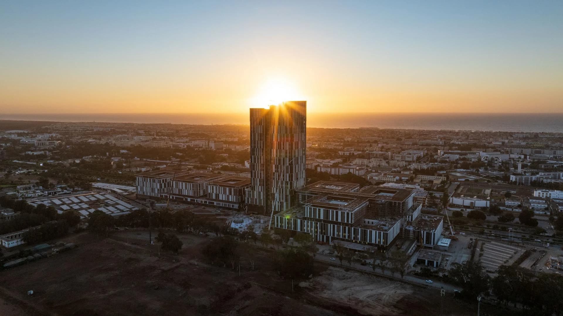 Vue aérienne d'un gratte-ciel moderne aux couleurs sombres, avec le soleil éclatant se couchant ou se levant derrière lui, illuminant une ville côtière s'étendant vers l'océan.