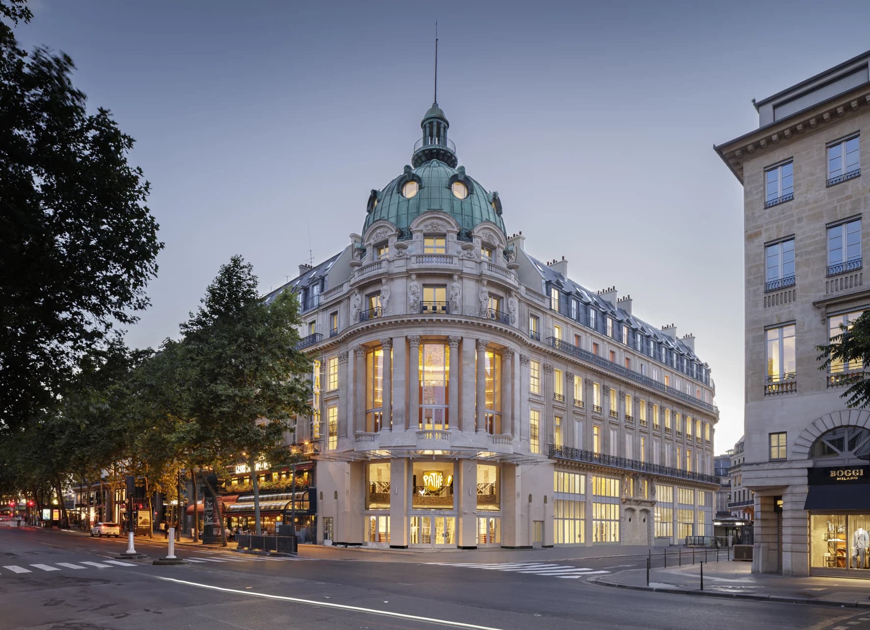 Un grand bâtiment parisien en pierre claire, orné d'un dôme vert-de-gris distinctif, est illuminé au crépuscule. Ses nombreuses fenêtres brillent de lumière chaude sur un carrefour urbain, avec le logo Pathé visible au rez-de-chaussée.