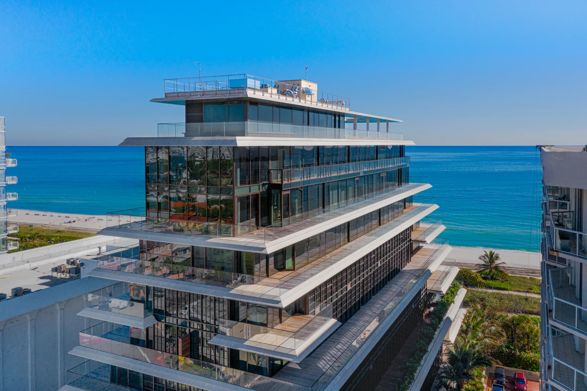 Immeuble moderne de grande hauteur avec une façade en verre réfléchissant et des balcons blancs en gradins donnant sur un océan bleu vif, une plage de sable blanc et un ciel clair.