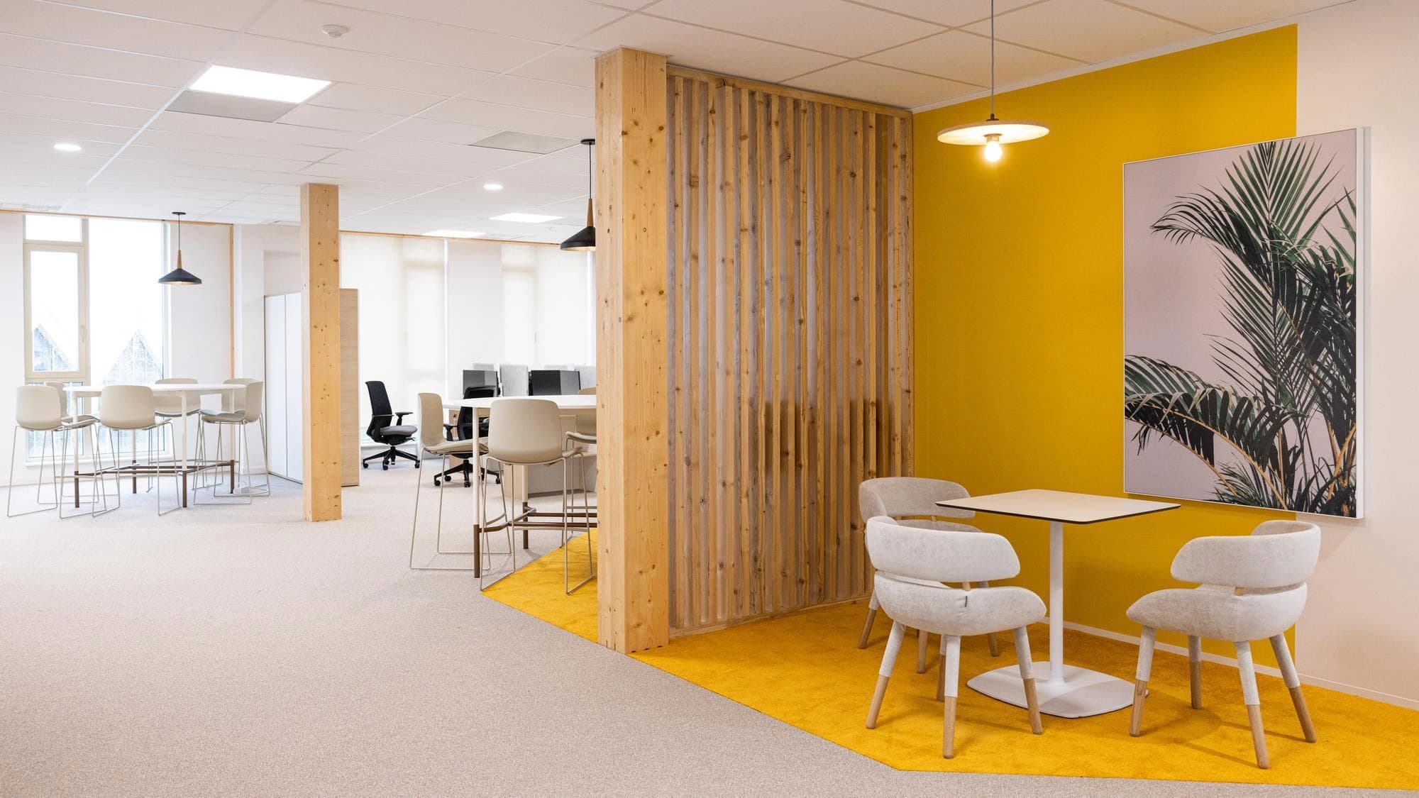 A bright, modern open-plan office features a light grey carpeted area with high tables and a wooden slatted partition leading to a vibrant yellow zone with a small table, light grey chairs, and a palm leaf artwork on the wall.