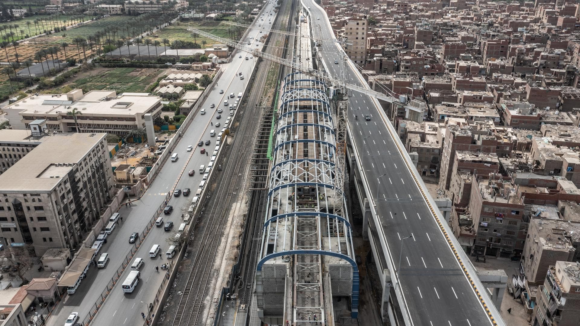 Vue aérienne d'une gare ferroviaire ou de métro moderne en construction, avec des arches bleues distinctives, située entre une autoroute existante et une nouvelle autoroute surélevée. Le paysage urbain dense présente des bâtiments en briques, des voies ferrées actives et des parcs de palmiers en arrière-plan.