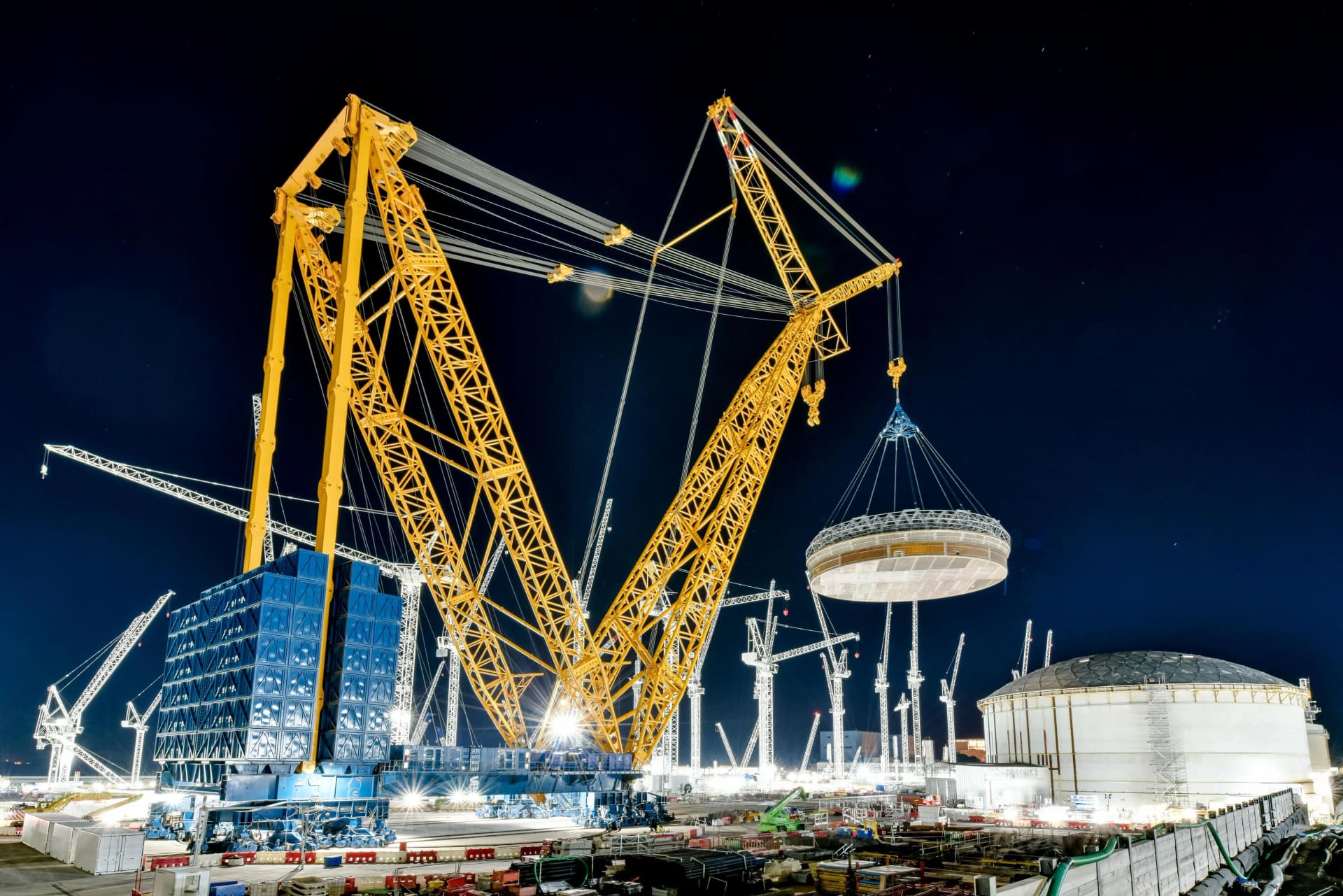 Une grue géante jaune soulève une grande structure circulaire blanche sur un chantier de construction éclairé de nuit, avec plusieurs autres grues et un réservoir en dôme visible à l'arrière-plan sous un ciel nocturne étoilé.
