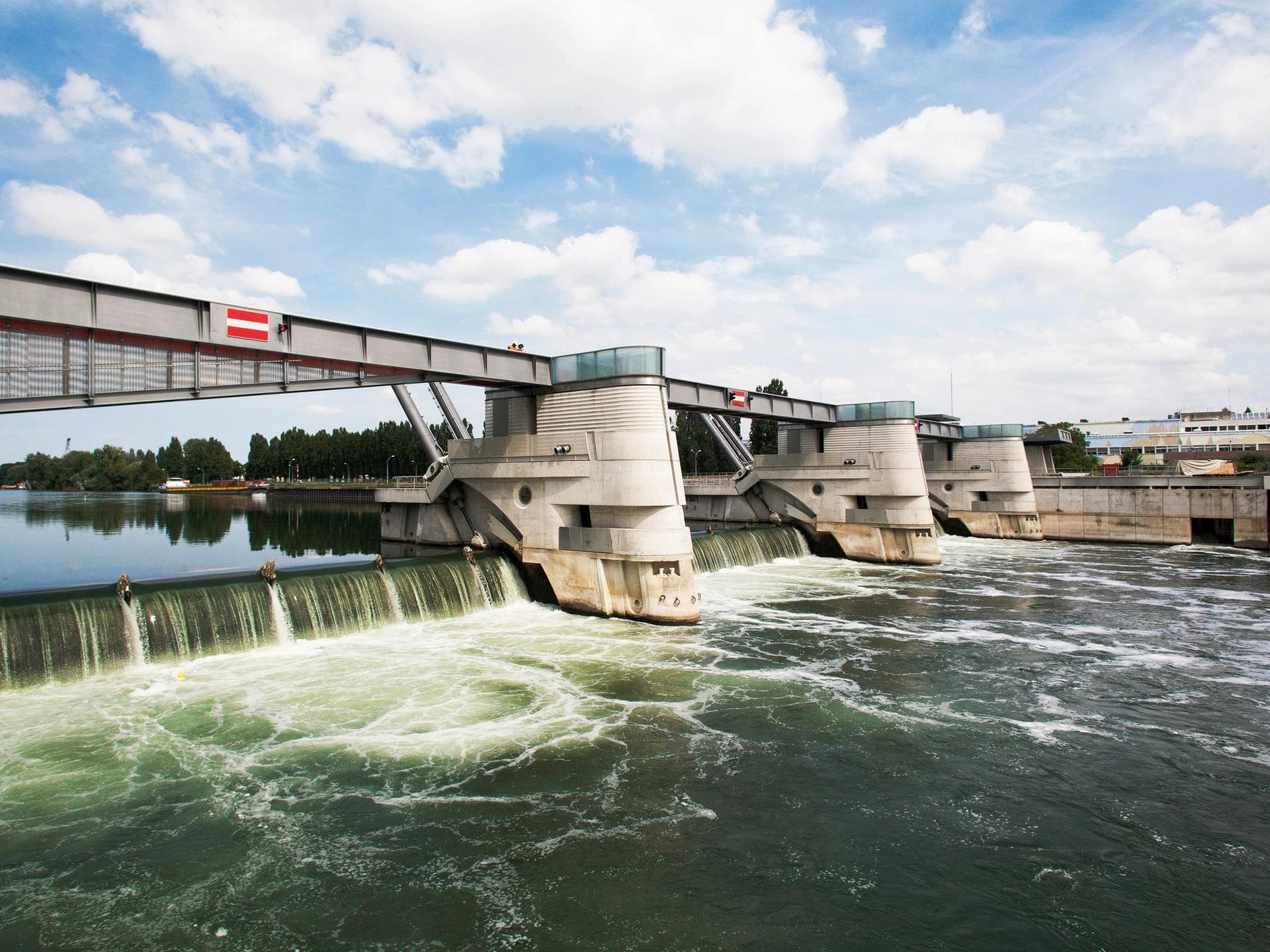 Un grand barrage moderne en béton avec une structure métallique, orné d'un drapeau rouge et blanc, enjambe une large rivière sous un ciel bleu parsemé de nuages blancs. L'eau cascade puissamment par plusieurs vannes, créant de l'écume blanche et des rapides verts et tumultueux en aval.