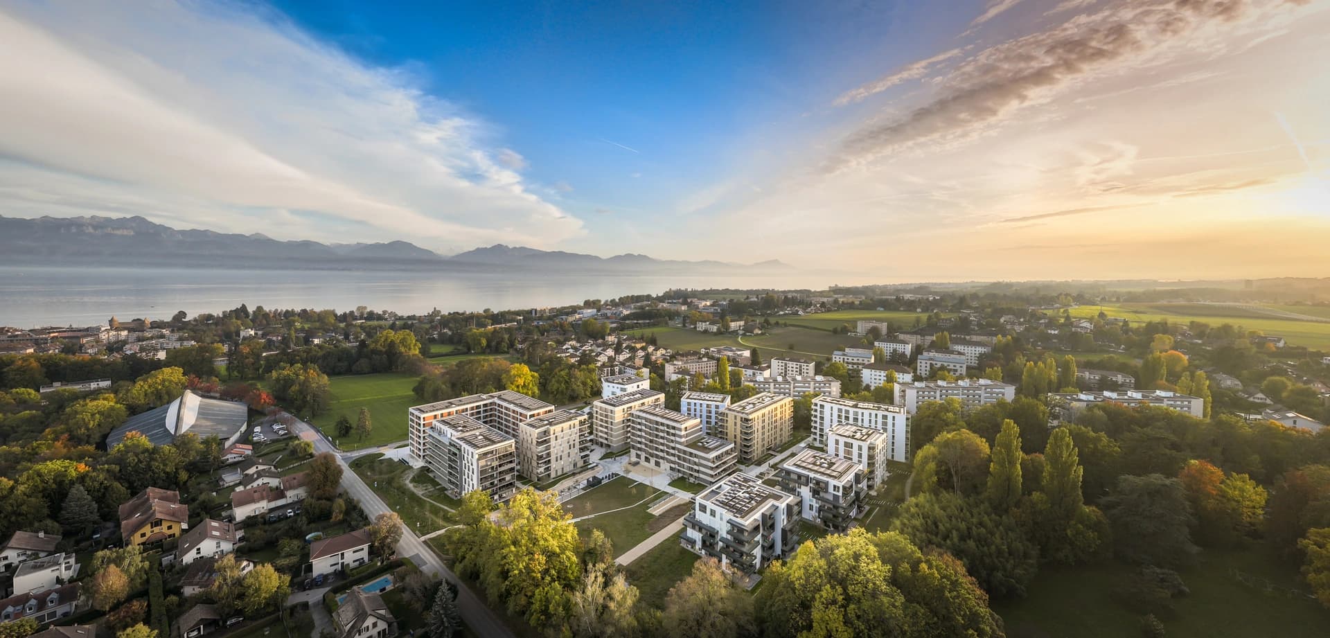 Vue aérienne d'un quartier moderne avec des immeubles clairs et des toits gris, entouré de verdure. Le panorama s'étend sur un grand lac, une ville et des montagnes à l'horizon, sous un ciel bleu clair baigné d'une lumière chaude de lever ou coucher de soleil.