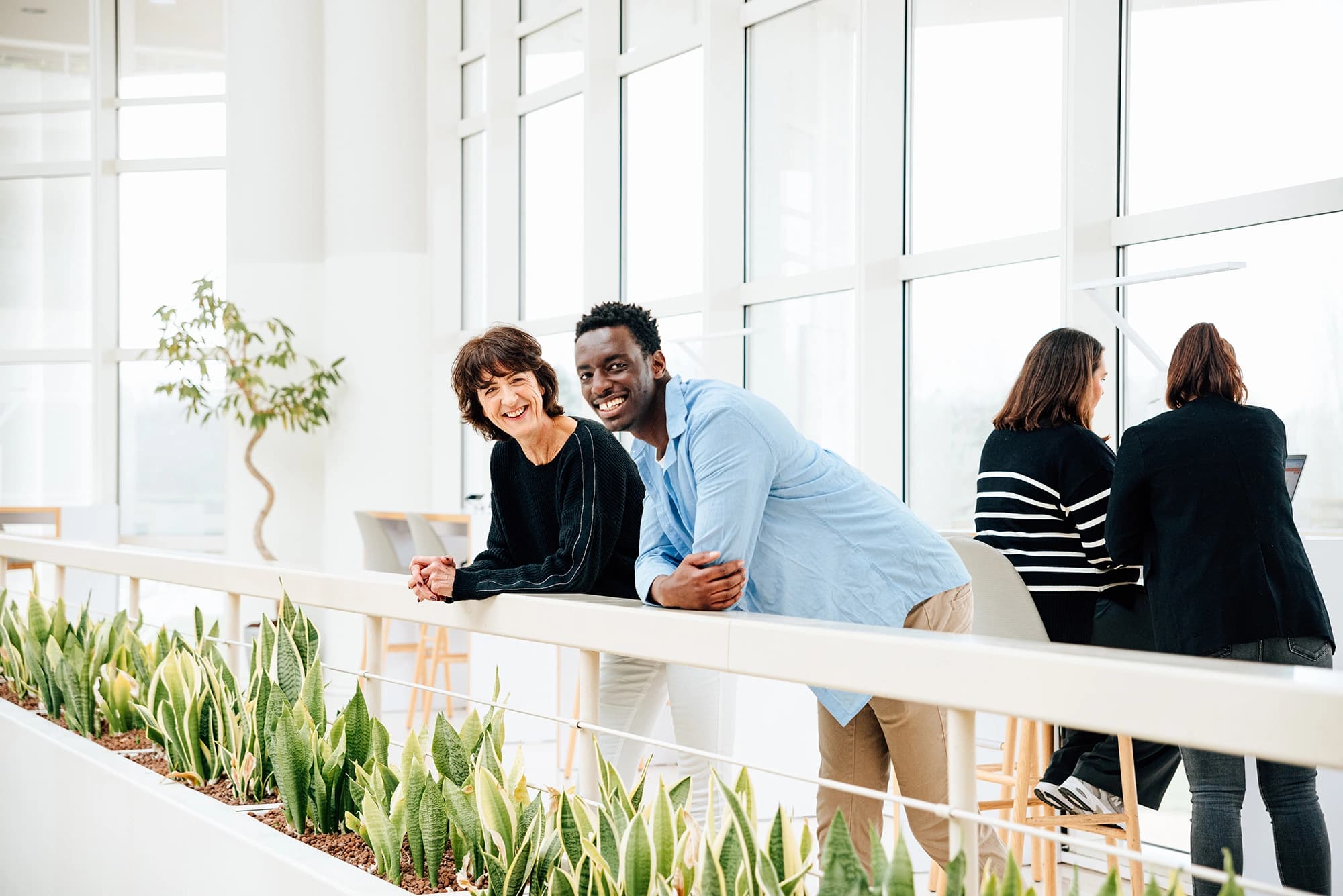 Un homme noir souriant en chemise bleue et une femme souriante en haut noir s'appuient sur une balustrade blanche avec des plantes vertes, dans un bureau moderne et lumineux. De grandes fenêtres et deux collègues travaillant sont visibles à l'arrière-plan.