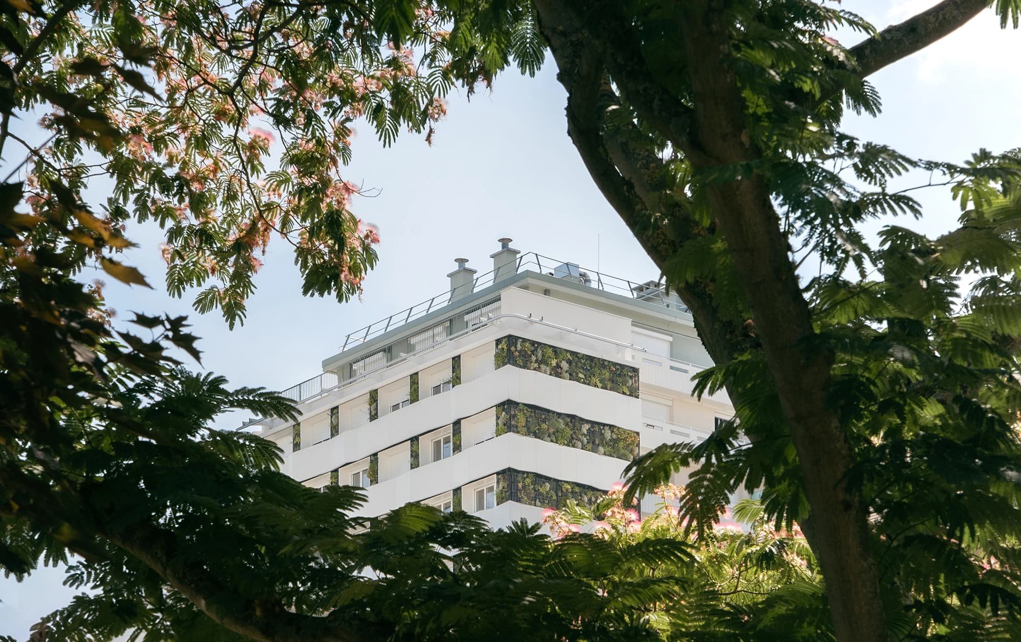 Un bâtiment moderne blanc avec des murs végétaux verts s'élève au-dessus des arbres verts luxuriants et des fleurs roses délicates, sous un ciel bleu clair.