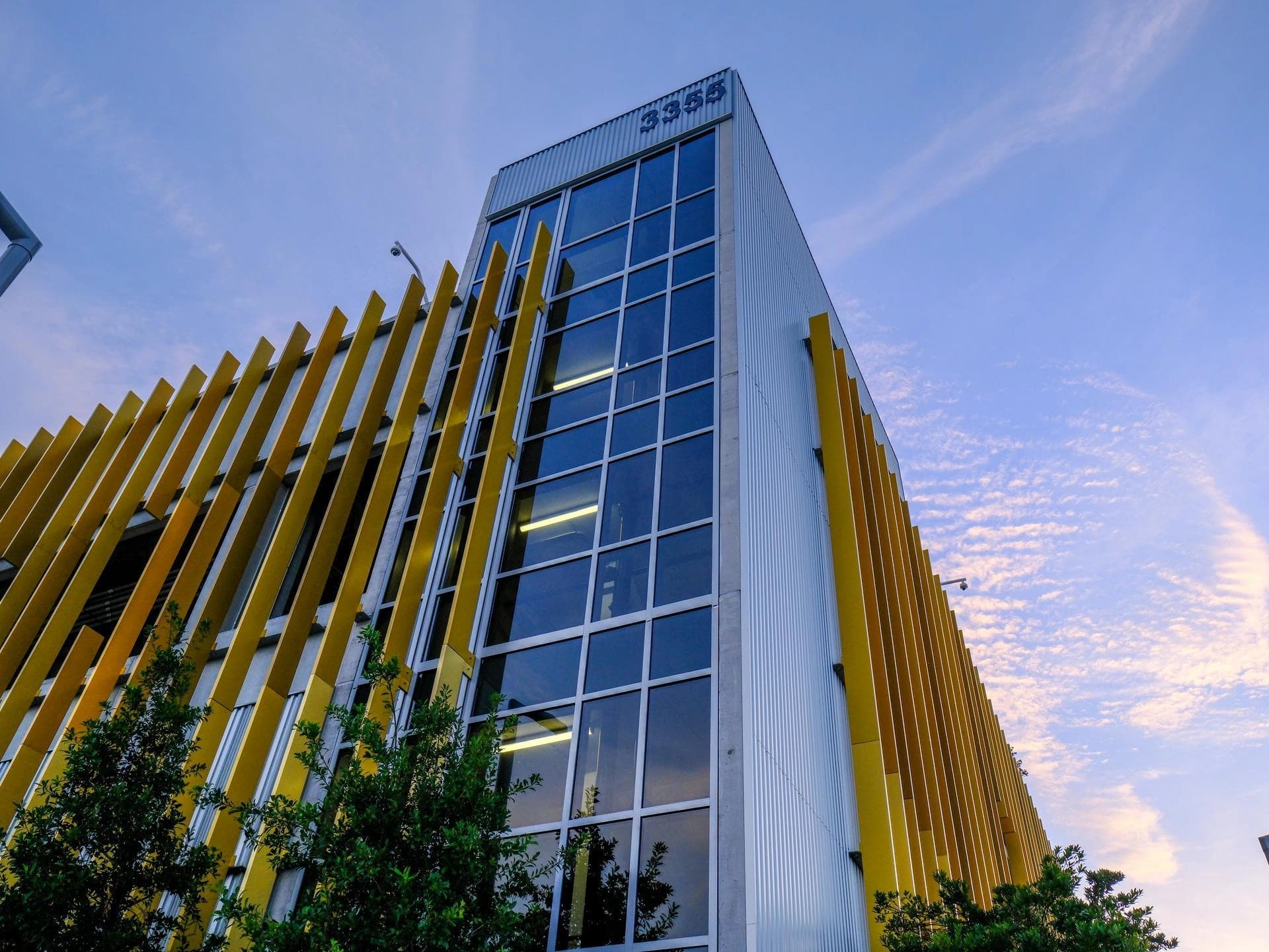 Un bâtiment moderne et angulaire, principalement gris métallique avec des persiennes verticales jaunes vives sur les côtés, présente une façade centrale en verre et porte le numéro 3355 au sommet, sous un ciel bleu-rose partiellement nuageux.