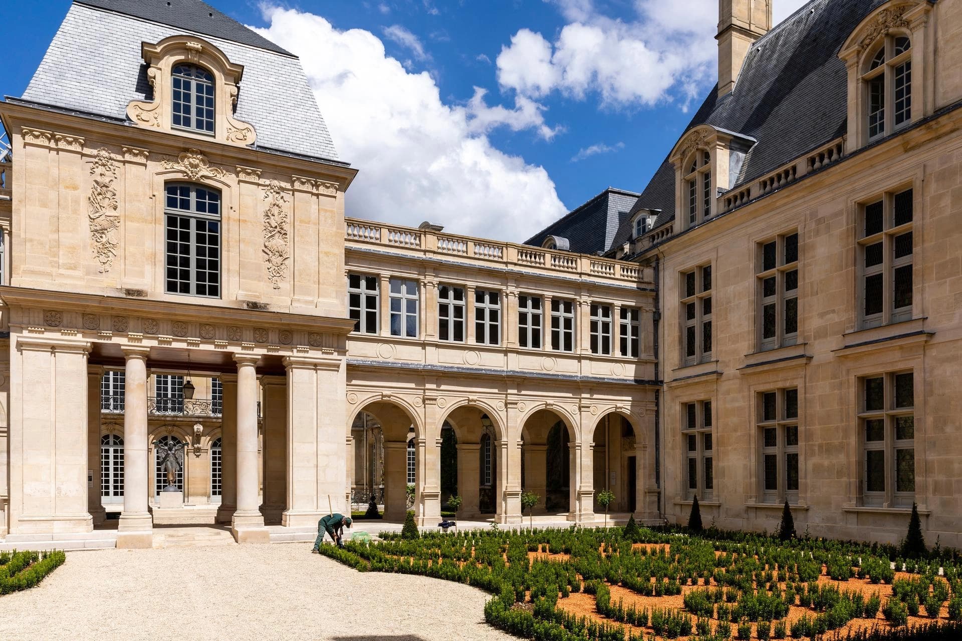 Un grand palais en pierre beige, avec des toits d'ardoise gris foncé et des fenêtres ornées, encadre une cour avec un jardin à la française. Un jardinier travaille parmi les buis verts taillés en motifs géométriques sur un parterre de gravier, sous un ciel bleu parsemé de nuages blancs.