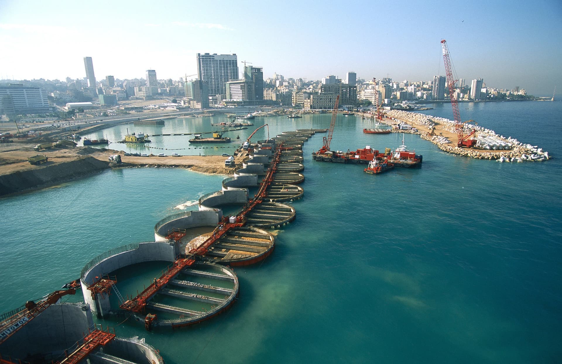 Une vue aérienne montre un vaste projet de construction côtière avec une ligne courbe de fondations circulaires en béton dans une eau bleu turquoise, flanquée de grues rouges en activité et d'un nouveau brise-lames avec des blocs de béton blancs. Une ligne d'horizon urbaine dense se dresse en arrière-plan sous un ciel clair.