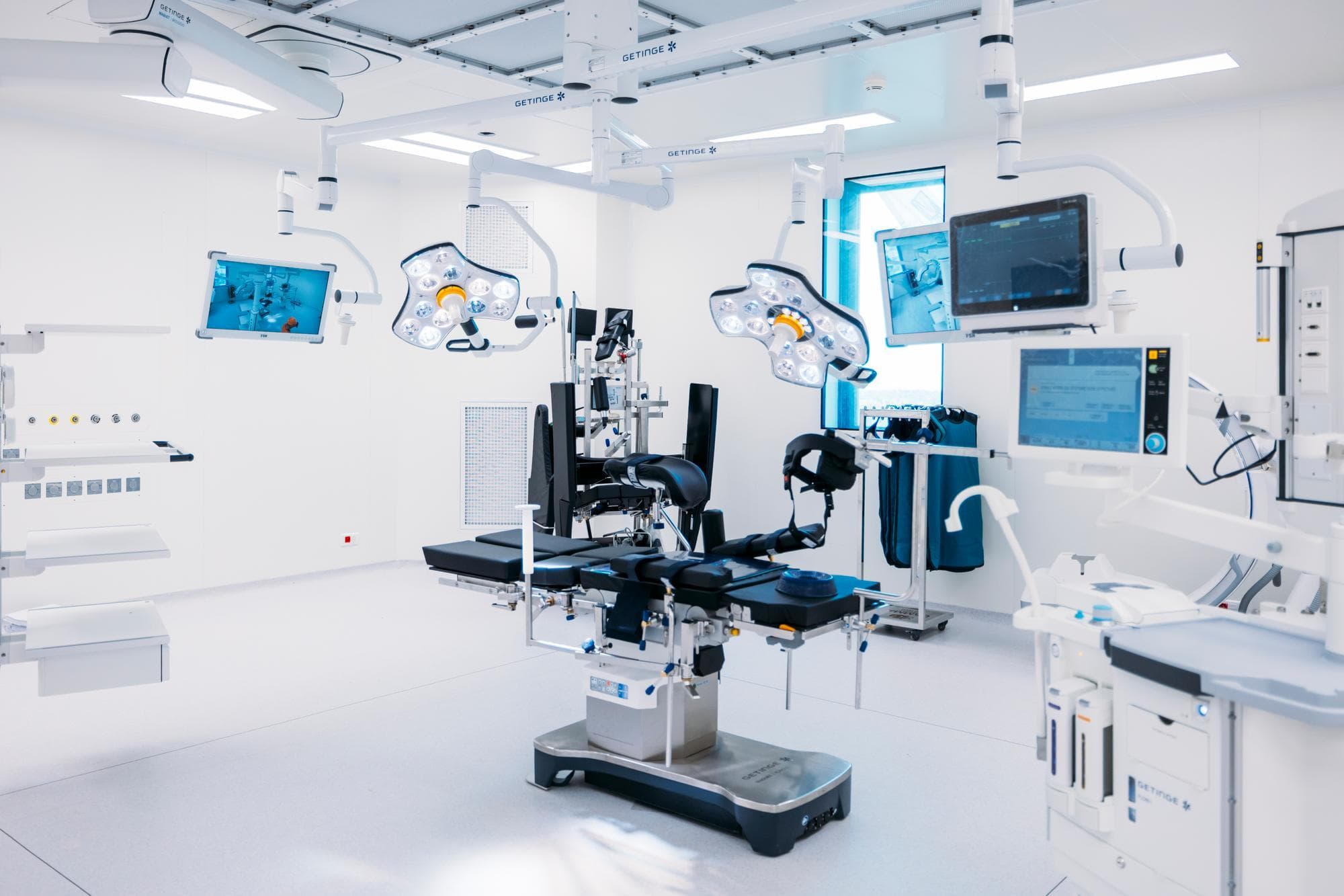 Une salle d'opération stérile et lumineuse est équipée d'une table chirurgicale centrale à rembourrage noir, de lampes opératoires suspendues et de nombreux moniteurs médicaux haute technologie, le tout dans des tons majoritairement blancs et gris clairs avec des accents bleus.