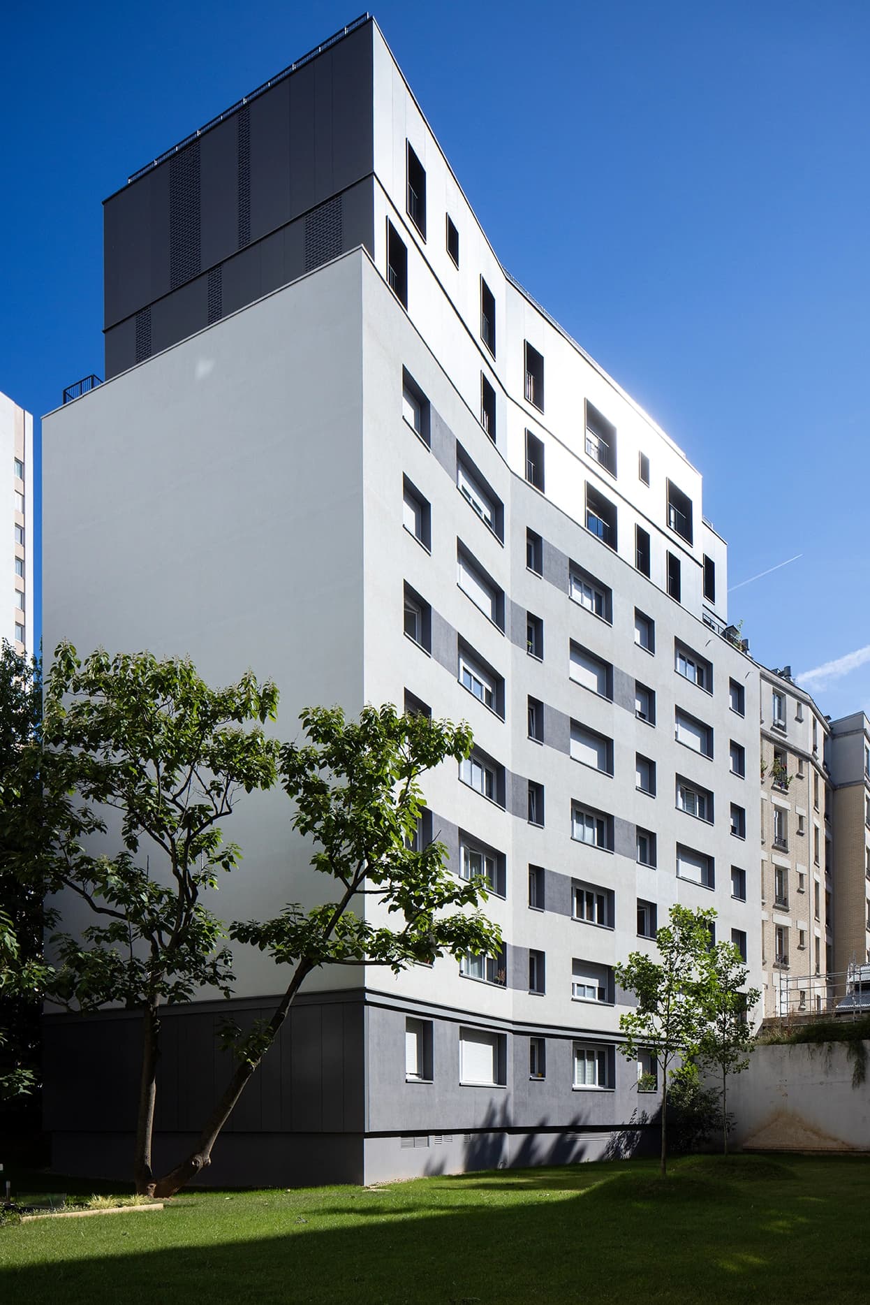 Un bâtiment résidentiel moderne de plusieurs étages, avec une façade incurvée en panneaux blancs et gris, se dresse sous un ciel bleu vif au-dessus d'une pelouse verte luxuriante avec des arbres.