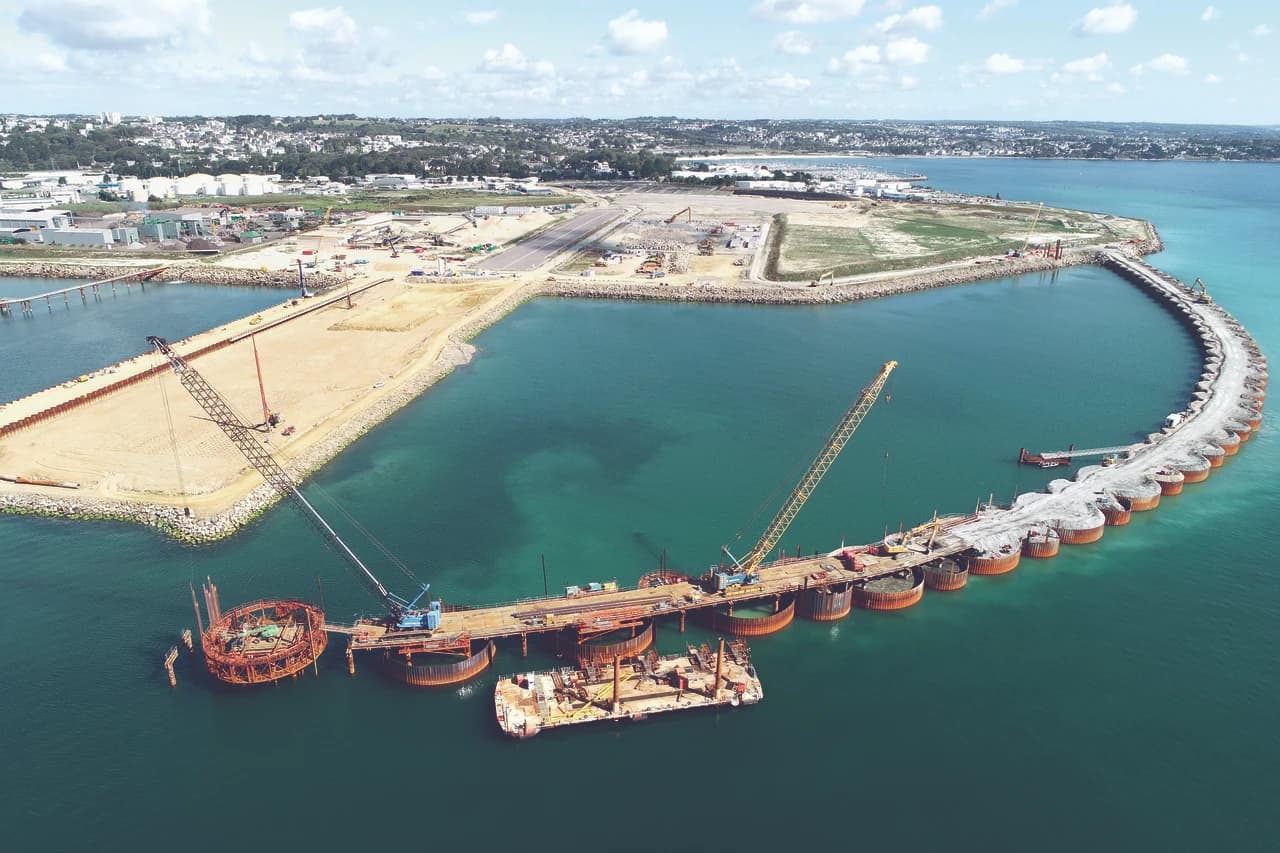 Vue aérienne d'un grand chantier de construction maritime, montrant des grues travaillant sur une longue digue incurvée faite de caissons dans une eau turquoise, et l'aménagement de terre-pleins côtiers avec des infrastructures urbaines et industrielles en arrière-plan.