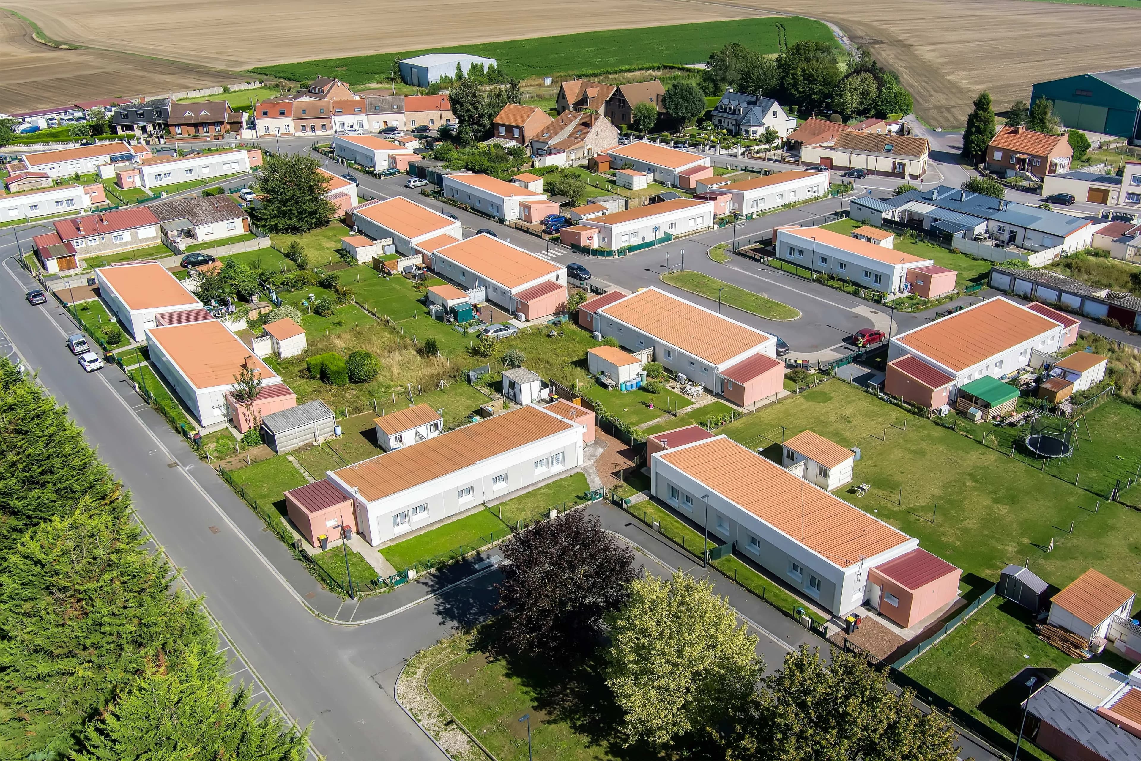 Vue aérienne d'un lotissement résidentiel moderne avec de nombreuses maisons rectangulaires aux murs clairs et toits orange, entourées de pelouses vertes et d'arbres, bordé par des champs agricoles bruns et verts à l'arrière-plan.