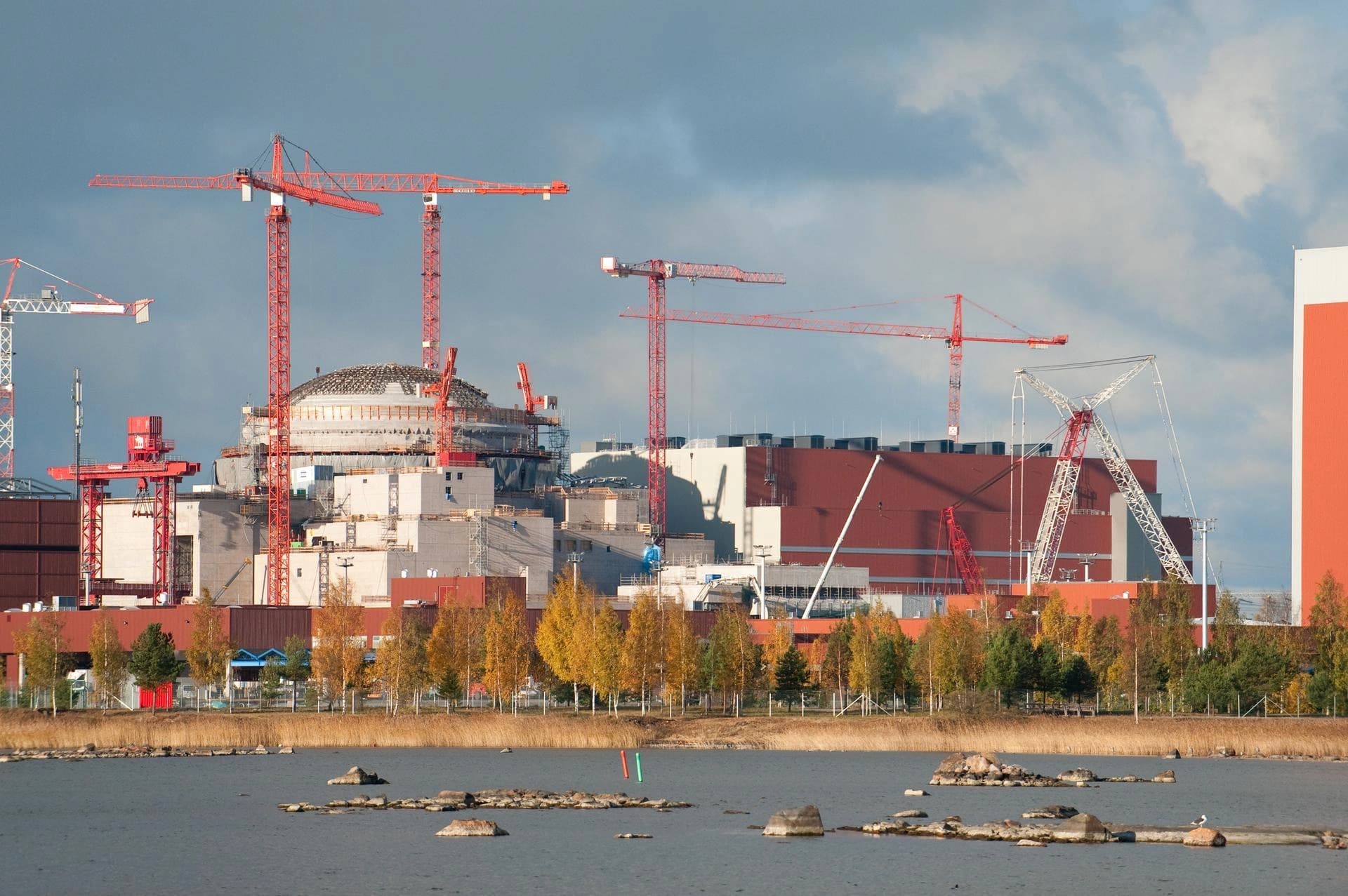 Vue d'un vaste site de construction industriel au bord de l'eau, avec de multiples grues rouges érigées autour d'un grand dôme et de bâtiments gris et marron. Des arbres aux couleurs automnales bordent la rive au premier plan, sous un ciel nuageux.
