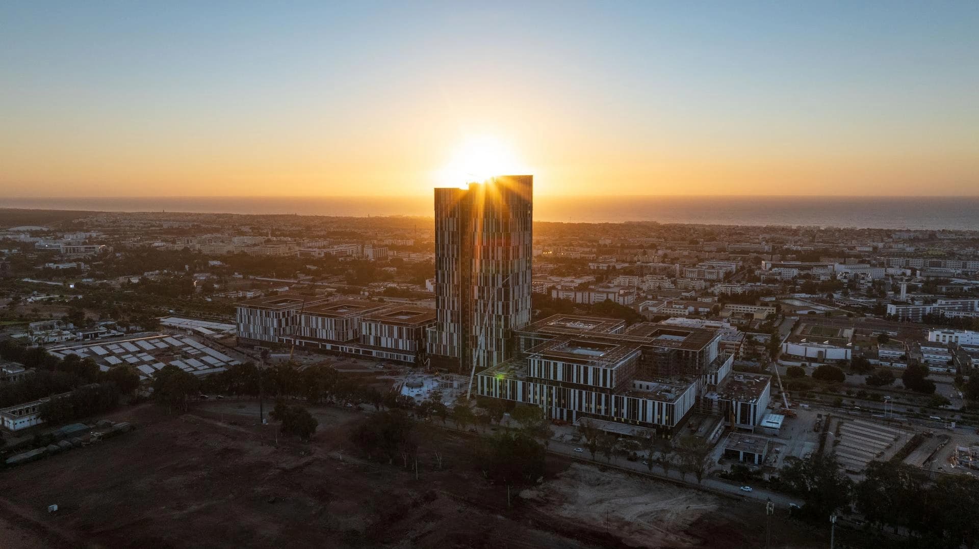 Vue aérienne d'un gratte-ciel moderne et de bâtiments adjacents, avec le soleil éclatant se couchant ou se levant juste derrière la tour. Le ciel affiche un dégradé d'orange et de bleu au-dessus d'une ville étendue longeant l'océan.
