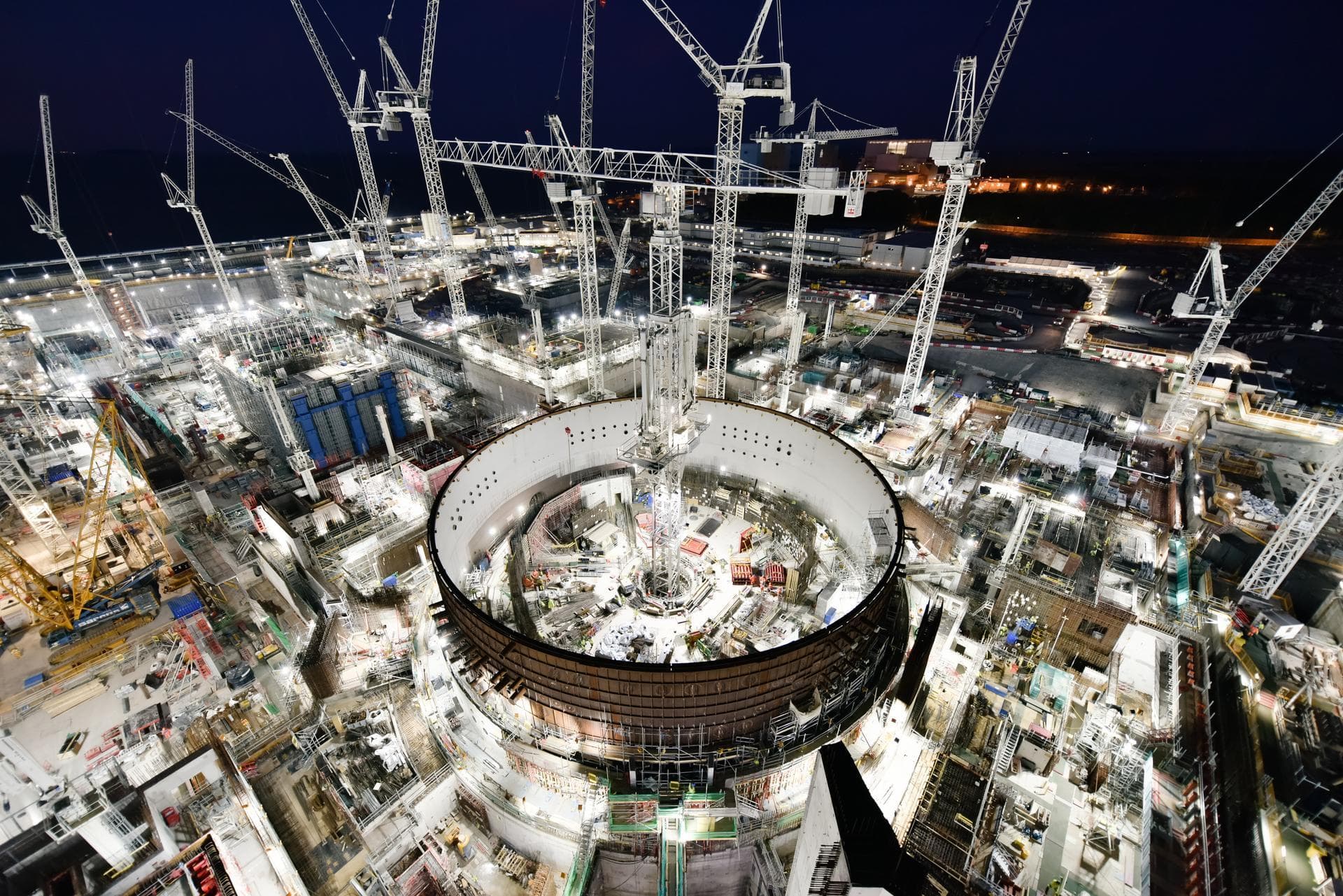 Vue aérienne nocturne d'un vaste chantier de construction industriel illuminé, avec de nombreuses grues blanches imposantes et une grande structure circulaire, potentiellement un réacteur de centrale nucléaire, en cours d'édification sous un ciel sombre.