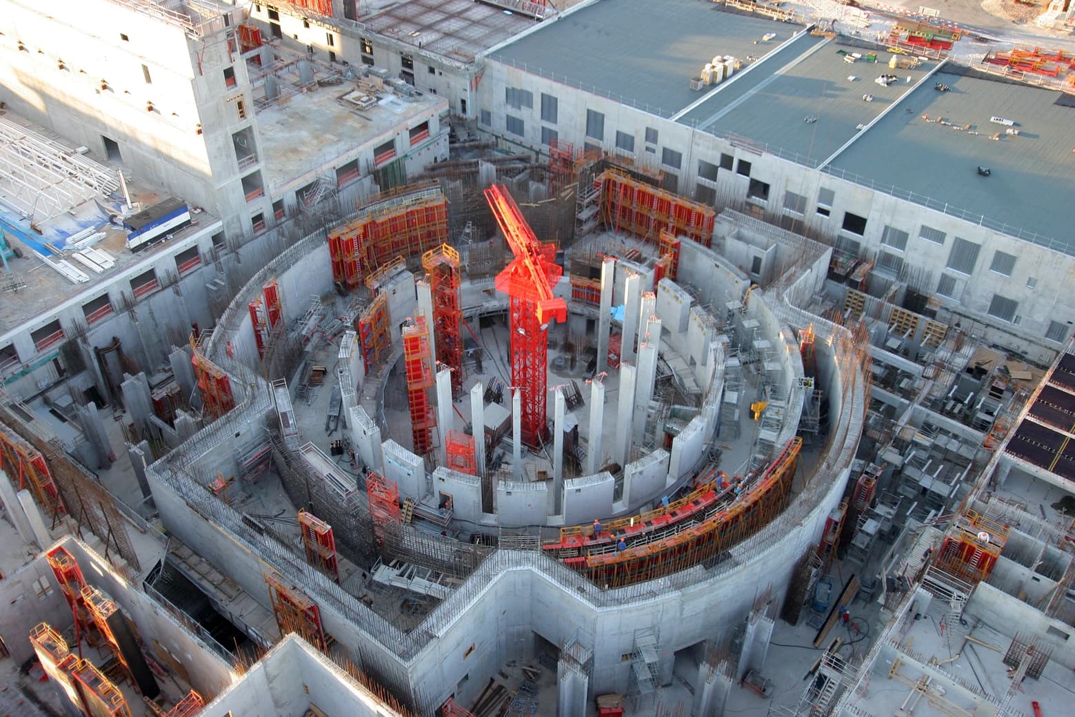Vue aérienne d'un vaste chantier de construction dominé par une grande structure circulaire en béton en cours, avec une grue rouge centrale et de nombreux coffrages orange vif. Des murs et piliers gris sont entourés d'échafaudages, indiquant une phase précoce de construction.