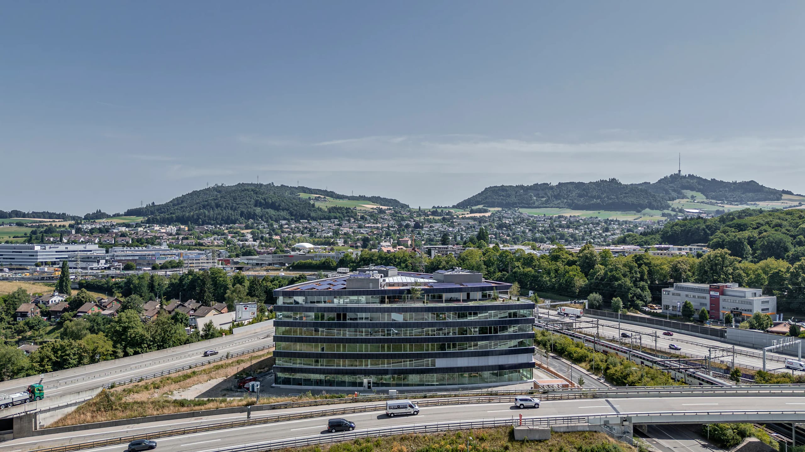 Une vue aérienne présente un immeuble de bureaux moderne avec des panneaux solaires sur le toit, entouré d'un vaste réseau autoroutier. L'arrière-plan montre une ville s'étendant vers des collines boisées sous un ciel bleu clair.