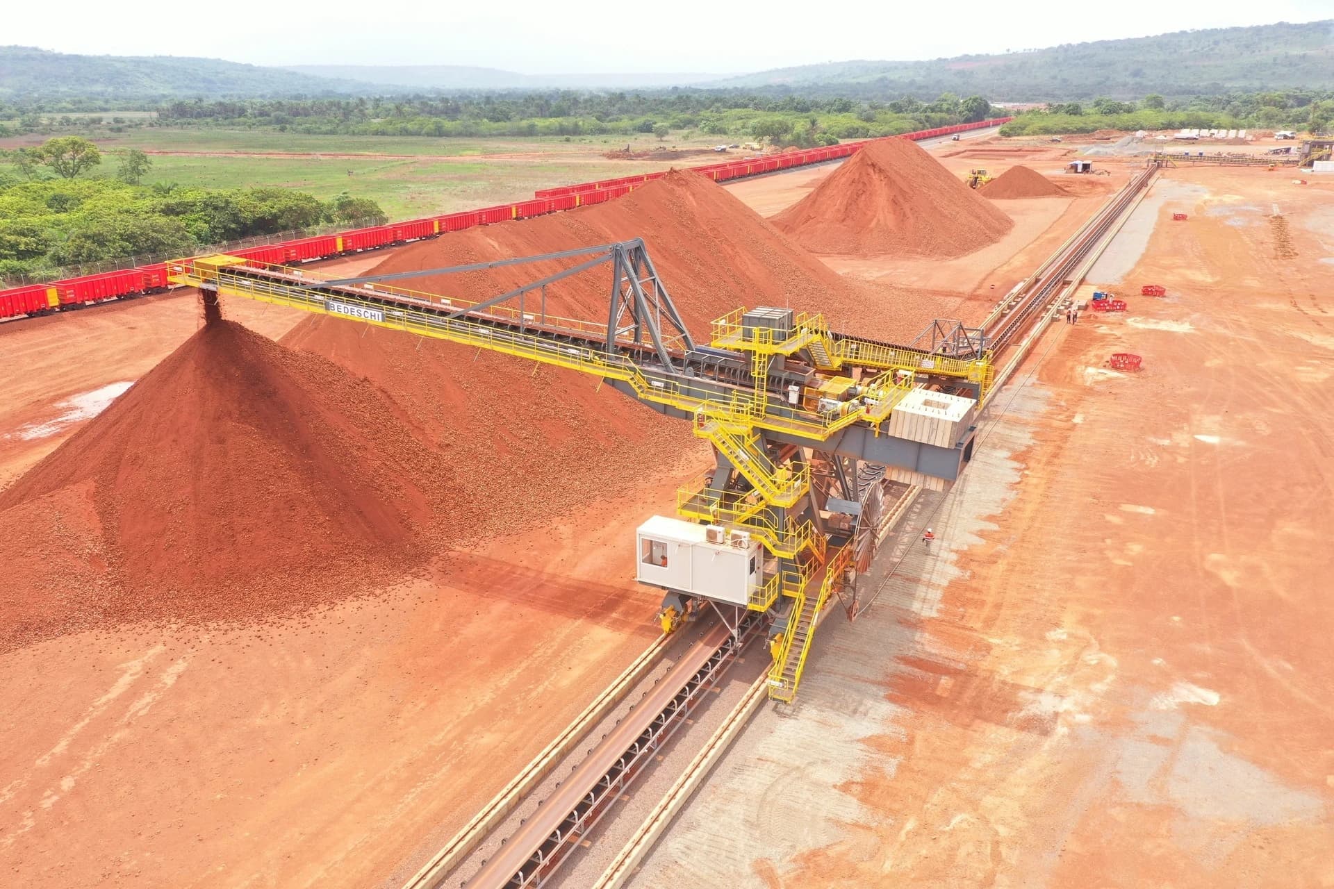 Vue aérienne d'une installation minière où un grand empileur jaune et gris dépose du minerai brun-rougeâtre sur d'immenses tas de stockage. Un long train de wagons rouges longe les tapis roulants et le sol orange poussiéreux, avec des collines verdoyantes à l'horizon.