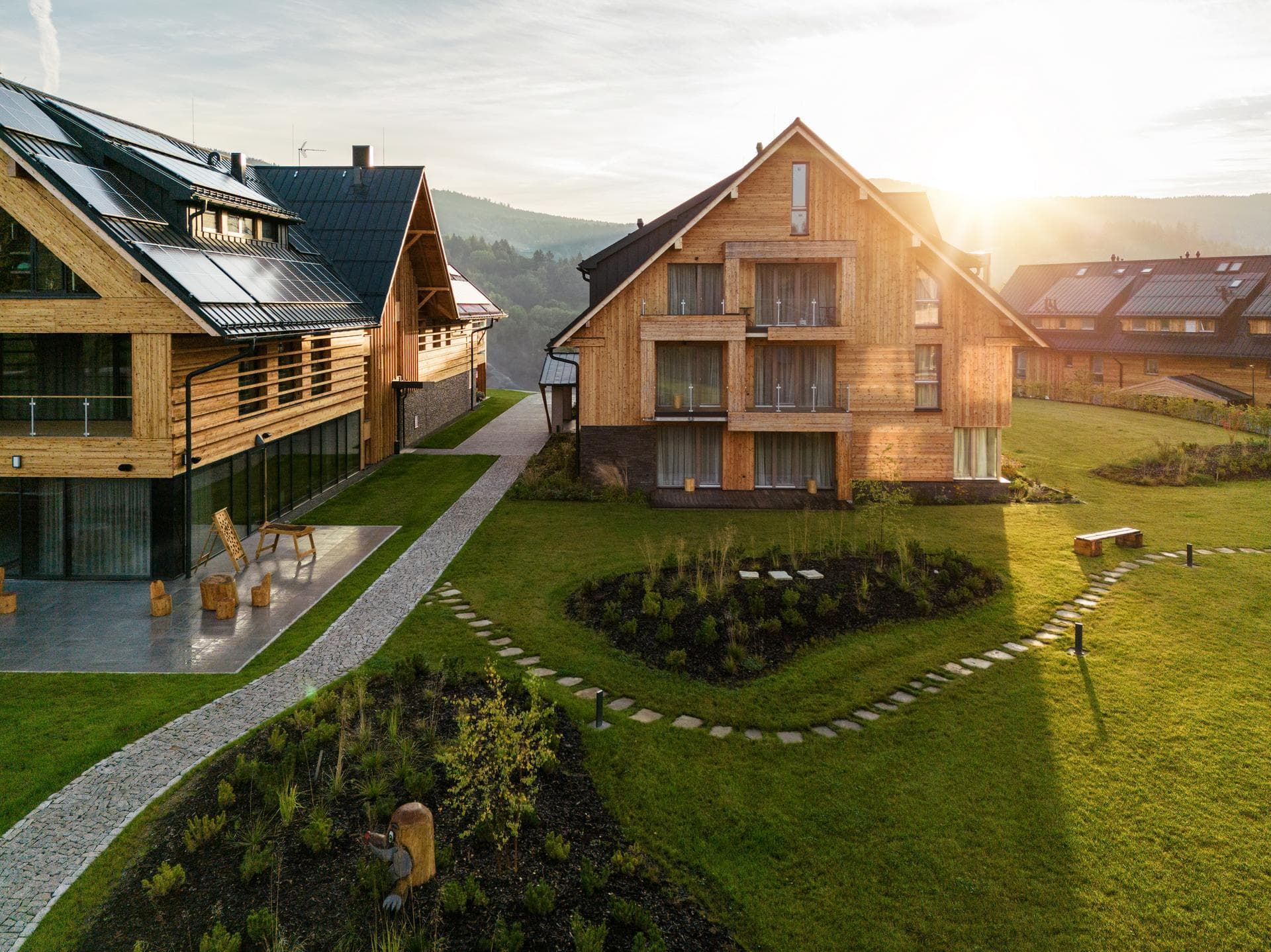 Deux bâtiments contemporains en bois avec des toits en pente, l'un doté de panneaux solaires et l'autre de multiples balcons, se dressent sur une pelouse verte luxuriante ponctuée de chemins en pierre. La scène est baignée par la lumière dorée et chaleureuse d'un soleil bas, avec des montagnes brumeuses en arrière-plan.