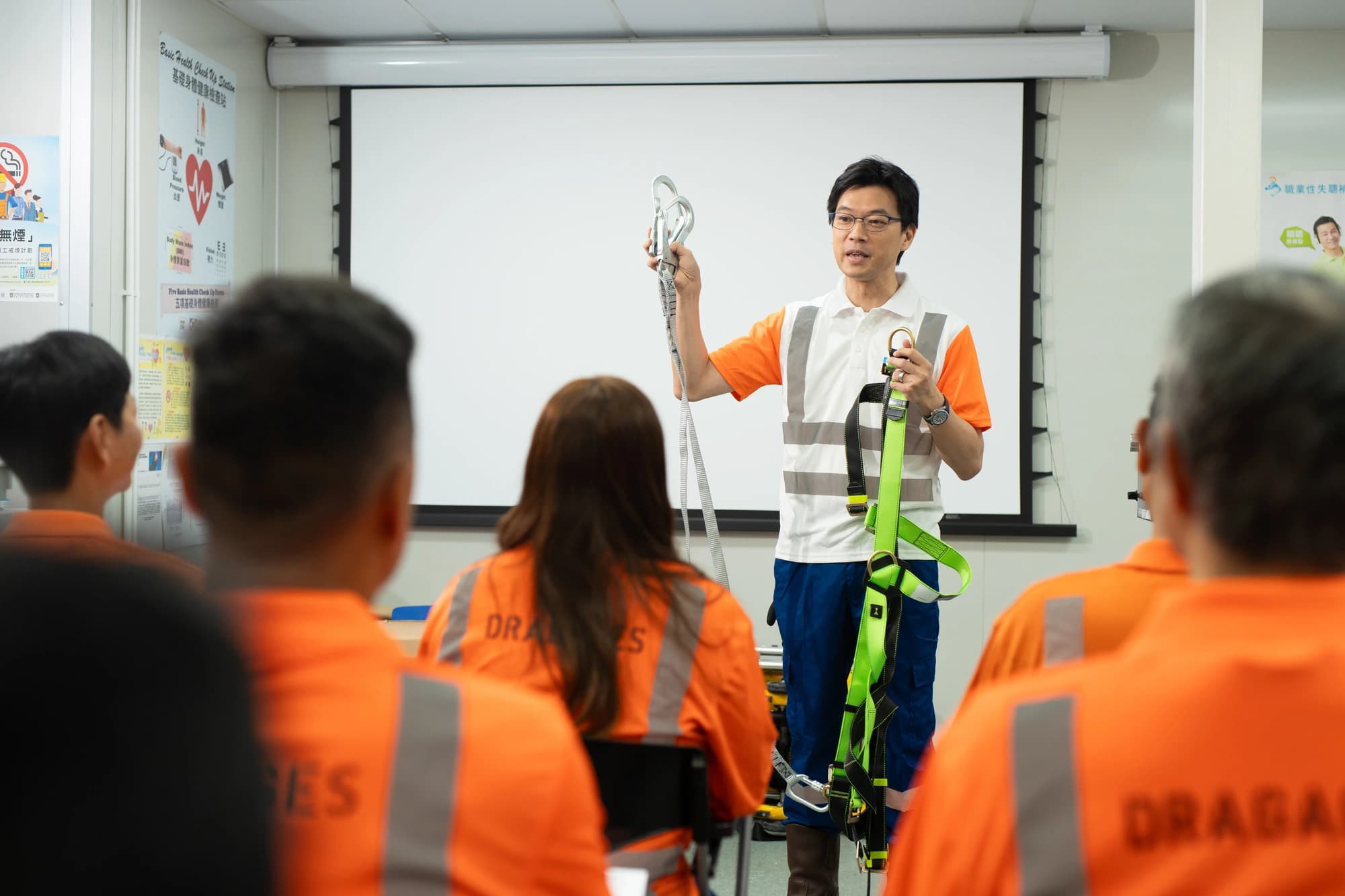 Un instructeur asiatique en tenue de sécurité orange et blanc présente un harnais et des mousquetons à une audience en uniforme de travail orange. La scène se déroule dans une salle de formation avec un écran de projection blanc à l'arrière-plan.
