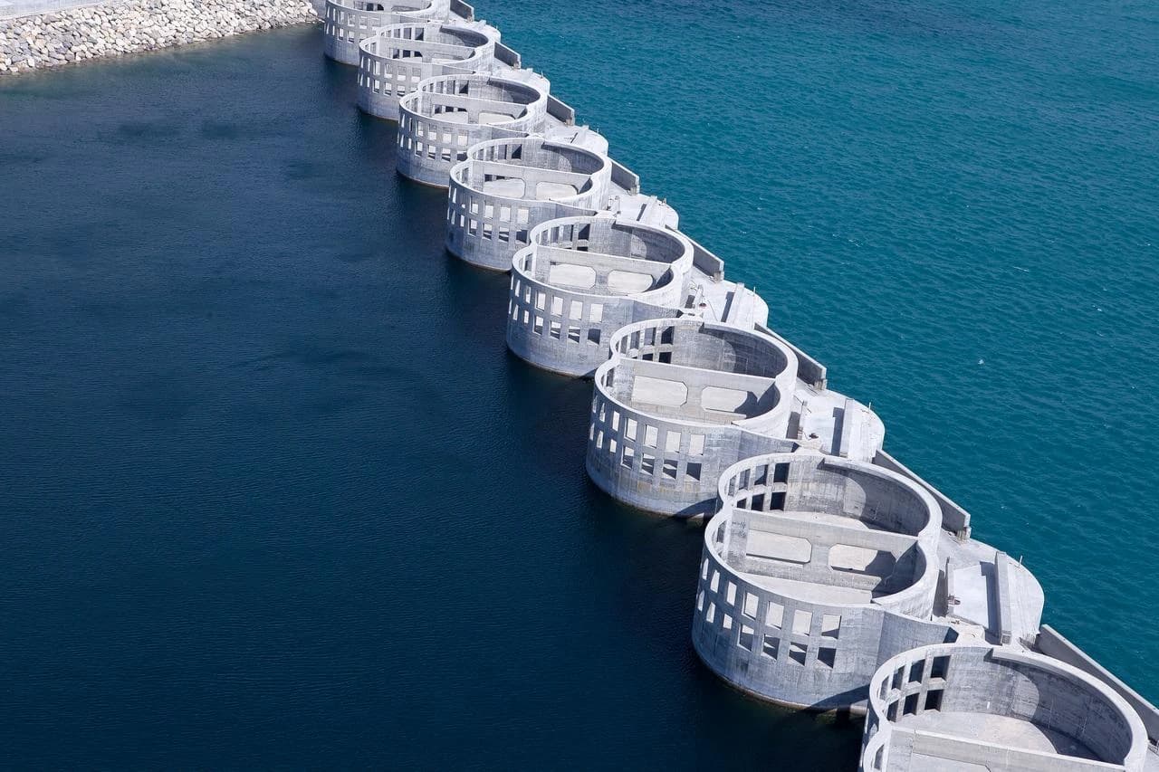 Vue aérienne d'une longue série de structures semi-circulaires en béton gris formant une barrière entre des eaux d'un bleu foncé et des eaux turquoise, suggérant un barrage ou un système de gestion hydraulique.