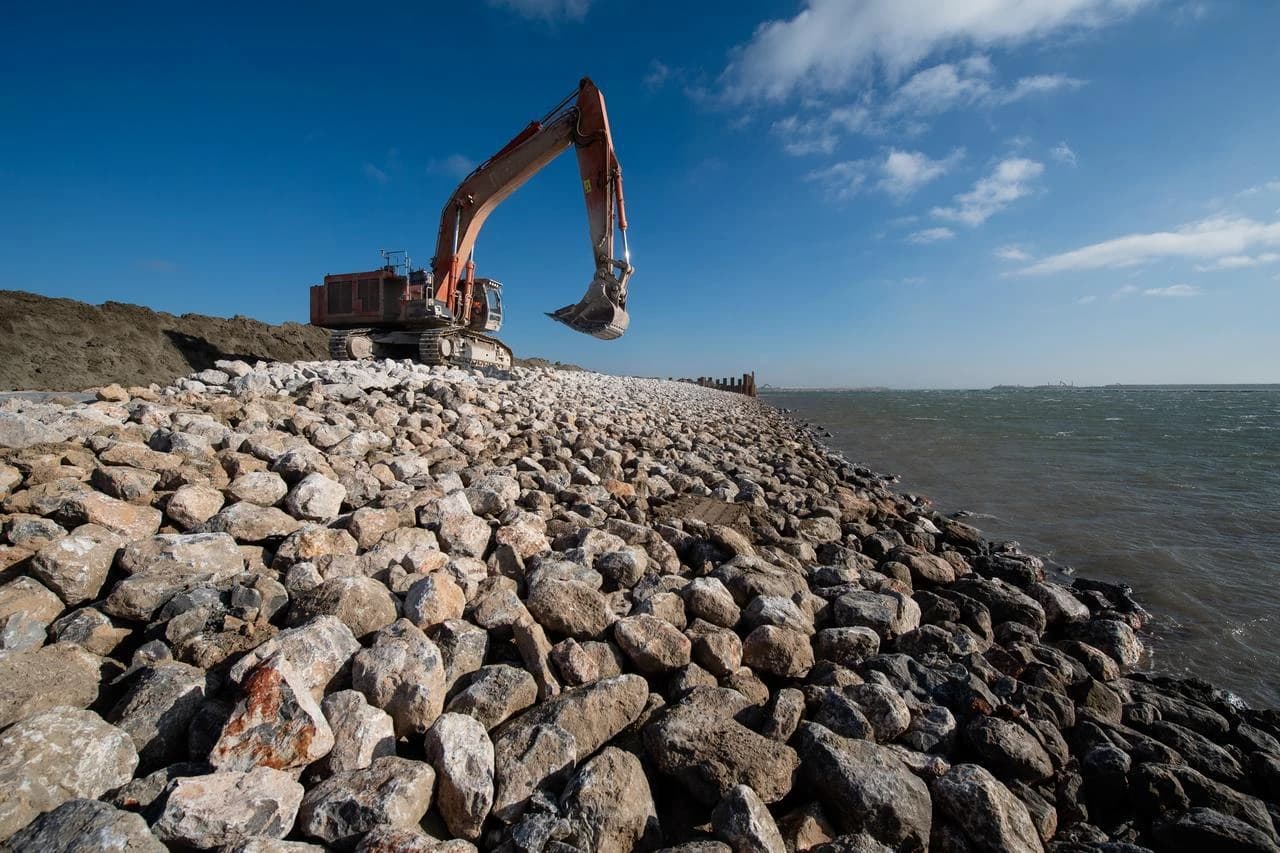 Un grand excavateur orange dépose des rochers sur une digue en construction longeant une mer agitée sous un ciel bleu.