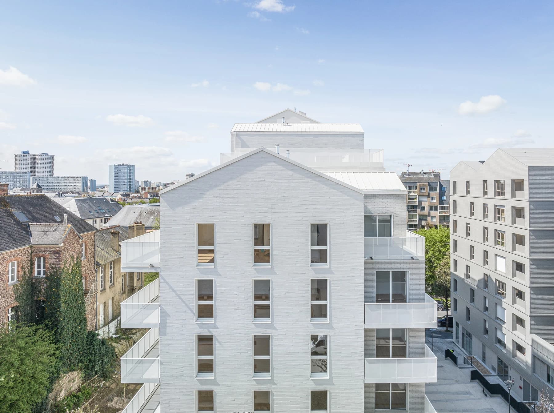 Vue d'ensemble de bâtiments résidentiels modernes blancs et gris avec des toits à pignon et des balcons, côtoyant d'anciennes maisons en pierre et un horizon urbain lointain sous un ciel bleu clair.