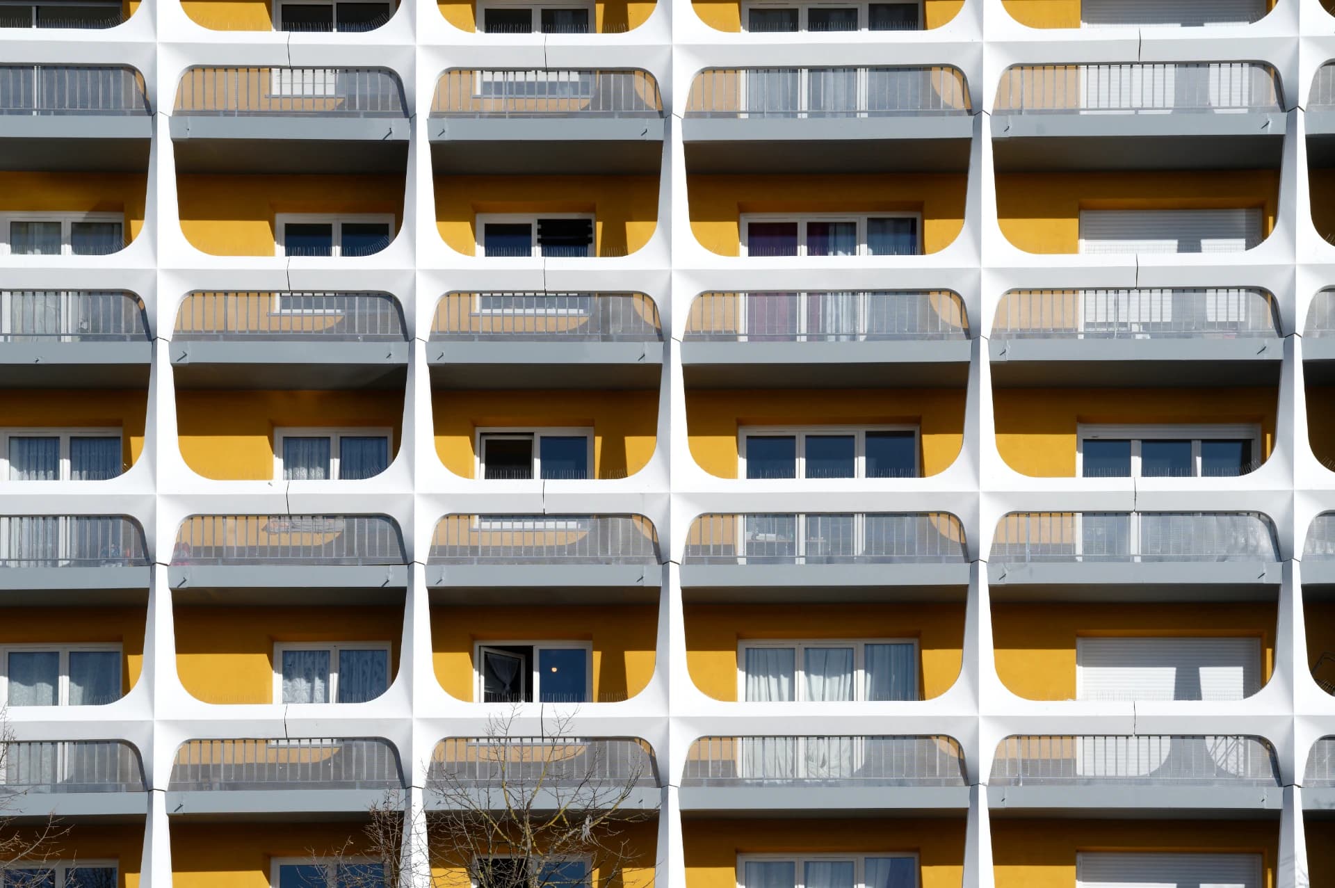 Façade d'immeuble moderne avec une grille répétitive de cadres architecturaux blancs arrondis encadrant des balcons et fenêtres aux murs jaunes vifs et garde-corps gris.