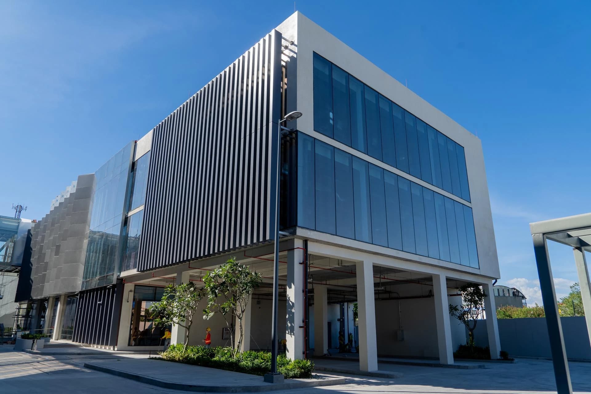 Bâtiment moderne blanc et gris avec des façades à lattes verticales noires et blanches et de grandes fenêtres en verre bleu. Son rez-de-chaussée est ouvert et soutenu par des piliers, sous un ciel bleu clair.