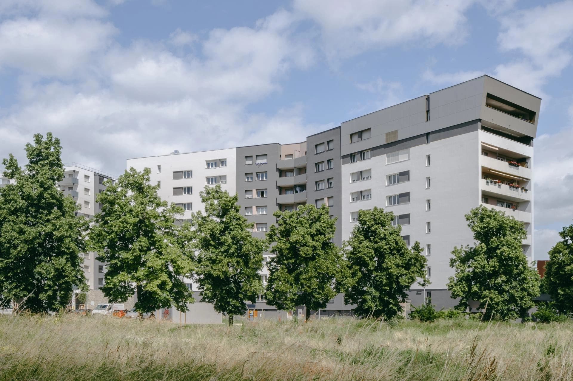 Complexe d'appartements moderne avec façades blanches et grises, rangées d'arbres et champ d'herbe sous un ciel bleu.