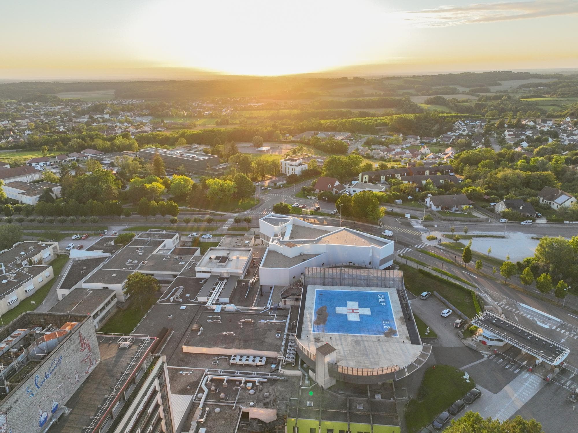Vue aérienne d'un complexe hospitalier moderne avec un héliport bleu et blanc sur le toit, entouré de zones résidentielles et de forêts verdoyantes. Le soleil couchant orange inonde le paysage d'une lumière chaude sous un ciel clair.
