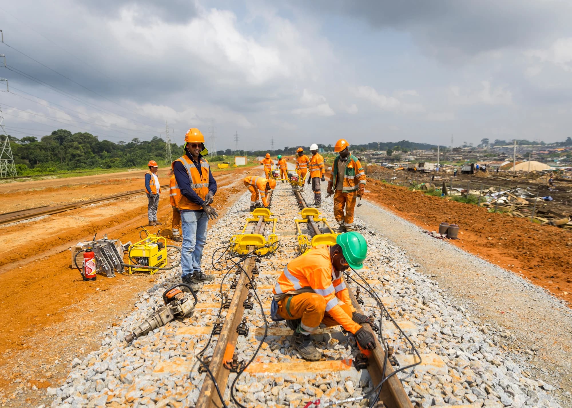 Plusieurs ouvriers en uniformes de sécurité orange et casques installent des voies ferrées sur un terrain de terre orange et de ballast, avec un ciel nuageux et des outils de chantier visibles.