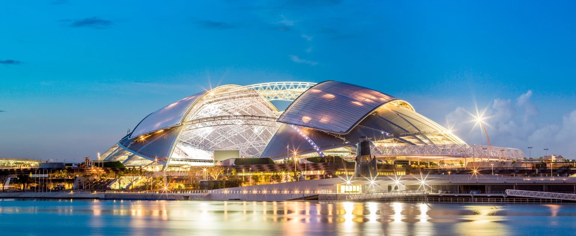 Vue nocturne du Singapore National Stadium, un stade moderne avec un toit dôme rétractable, intensément éclairé et se reflétant dans l'eau sous un ciel bleu crépusculaire.