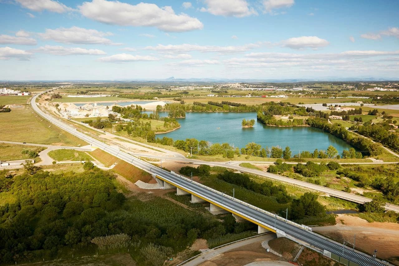 Vue aérienne d'une nouvelle ligne de chemin de fer sur un viaduc jaune et gris traversant un paysage verdoyant avec un grand lac turquoise, plusieurs routes et des constructions industrielles et urbaines en arrière-plan sous un ciel bleu nuageux.