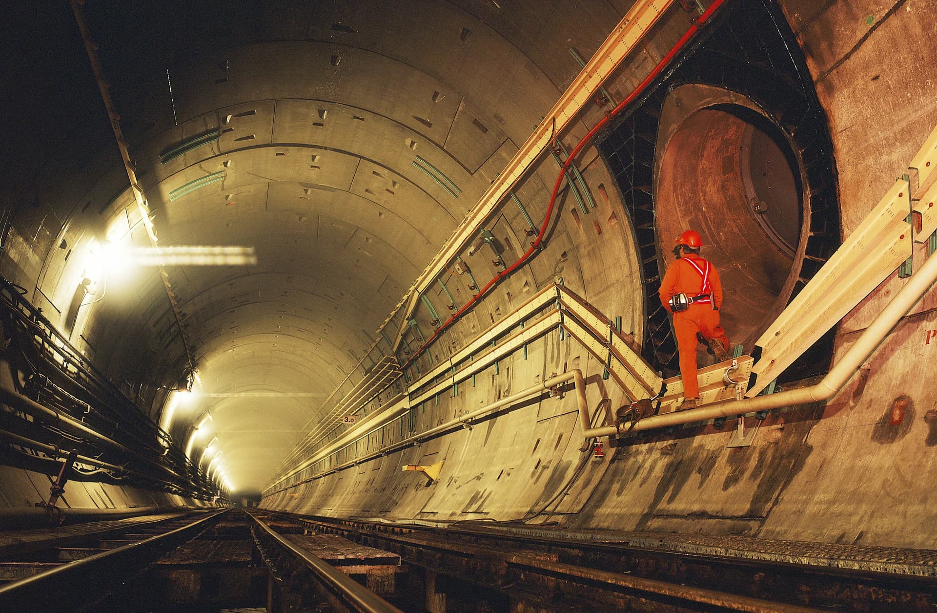 Un ouvrier en salopette orange et casque rouge se tient sur une passerelle latérale dans un long tunnel de béton éclairé par des lumières chaudes, avec des voies ferrées au sol et une grande ouverture cylindrique à droite.