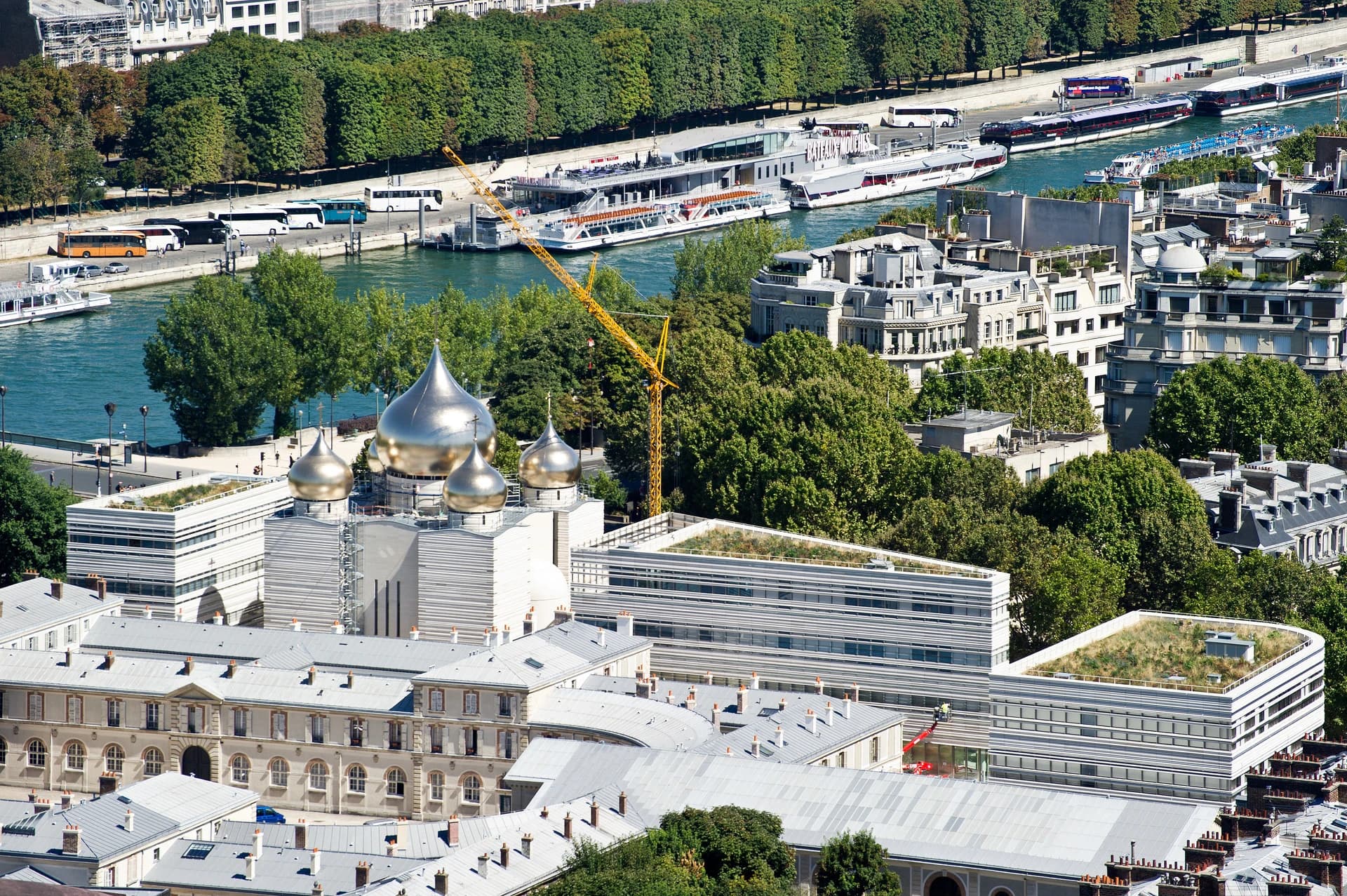 Vue aérienne du Centre orthodoxe russe à Paris avec ses dômes argentés et dorés, surplombant la Seine et ses bateaux amarrés, entouré d'immeubles classiques et de verdure sous un ciel ensoleillé.