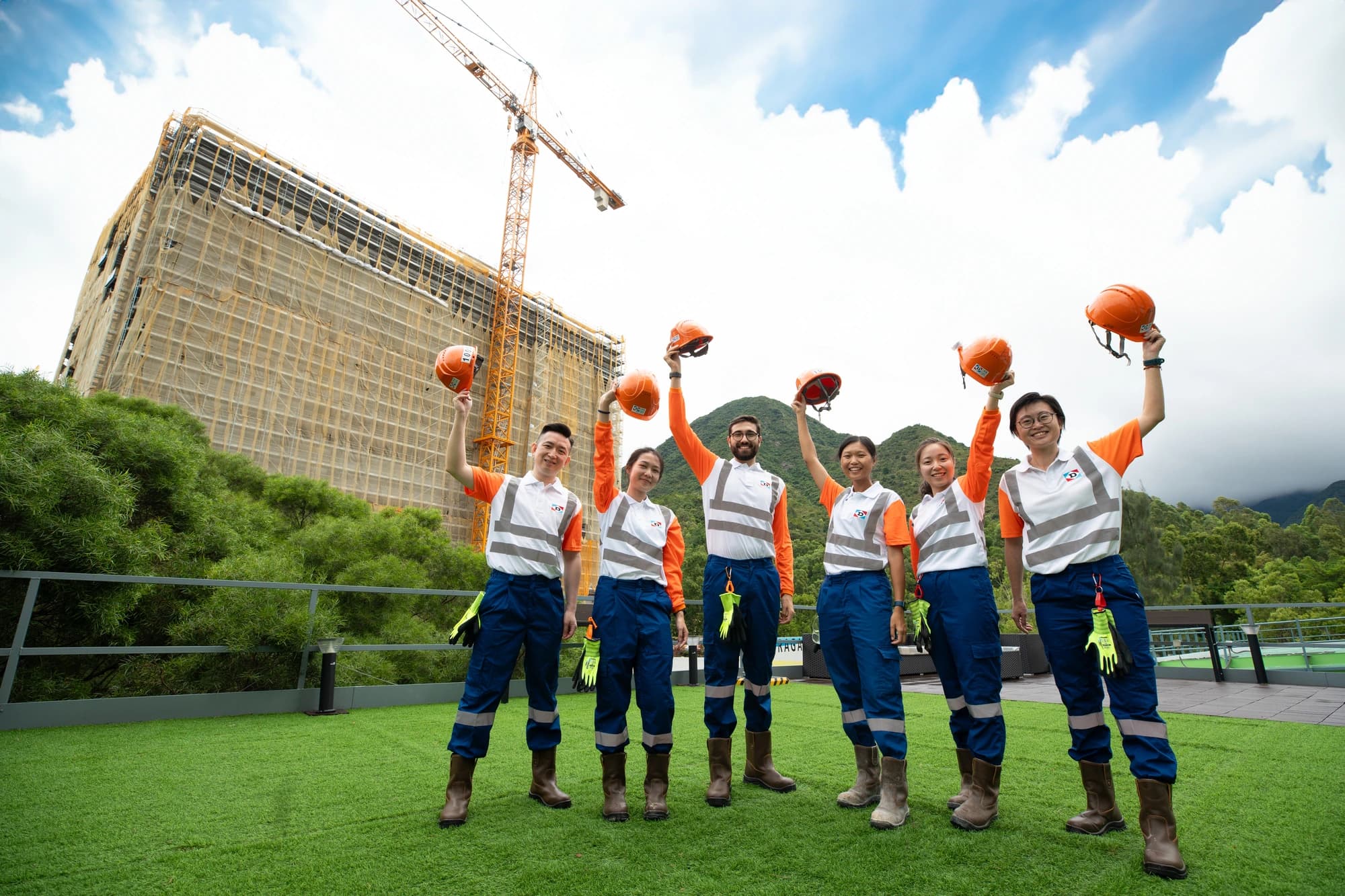 Six professionnels de la construction en uniformes de sécurité orange et blanc lèvent joyeusement leurs casques sur un toit en gazon synthétique. Derrière eux se dressent un grand bâtiment en construction avec des échafaudages et une grue, sous un ciel partiellement nuageux et des montagnes verdoyantes.