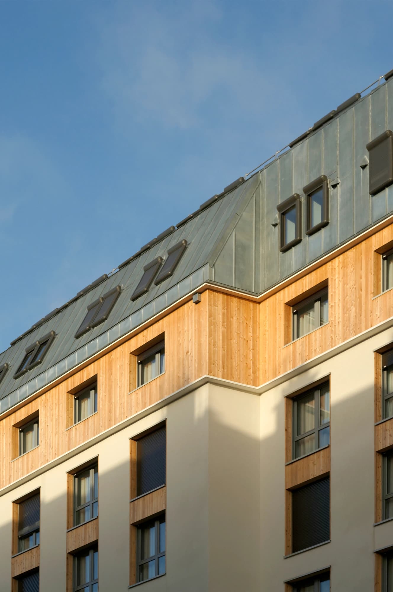 Vue architecturale d'un bâtiment moderne présentant un toit en zinc gris incliné avec des fenêtres de toit, un niveau supérieur en bardage de bois clair et une façade en enduit beige avec des fenêtres et des volets roulants sombres, sous un ciel bleu clair.
