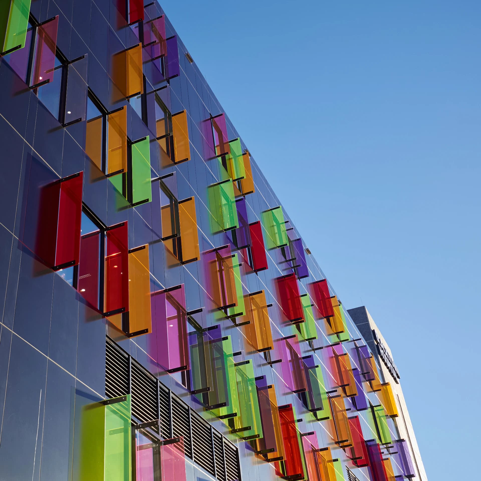 Façade d'un bâtiment moderne avec une grille de panneaux rectangulaires translucides et colorés (rouge, orange, vert, violet) sur un mur sombre, sous un ciel bleu clair.