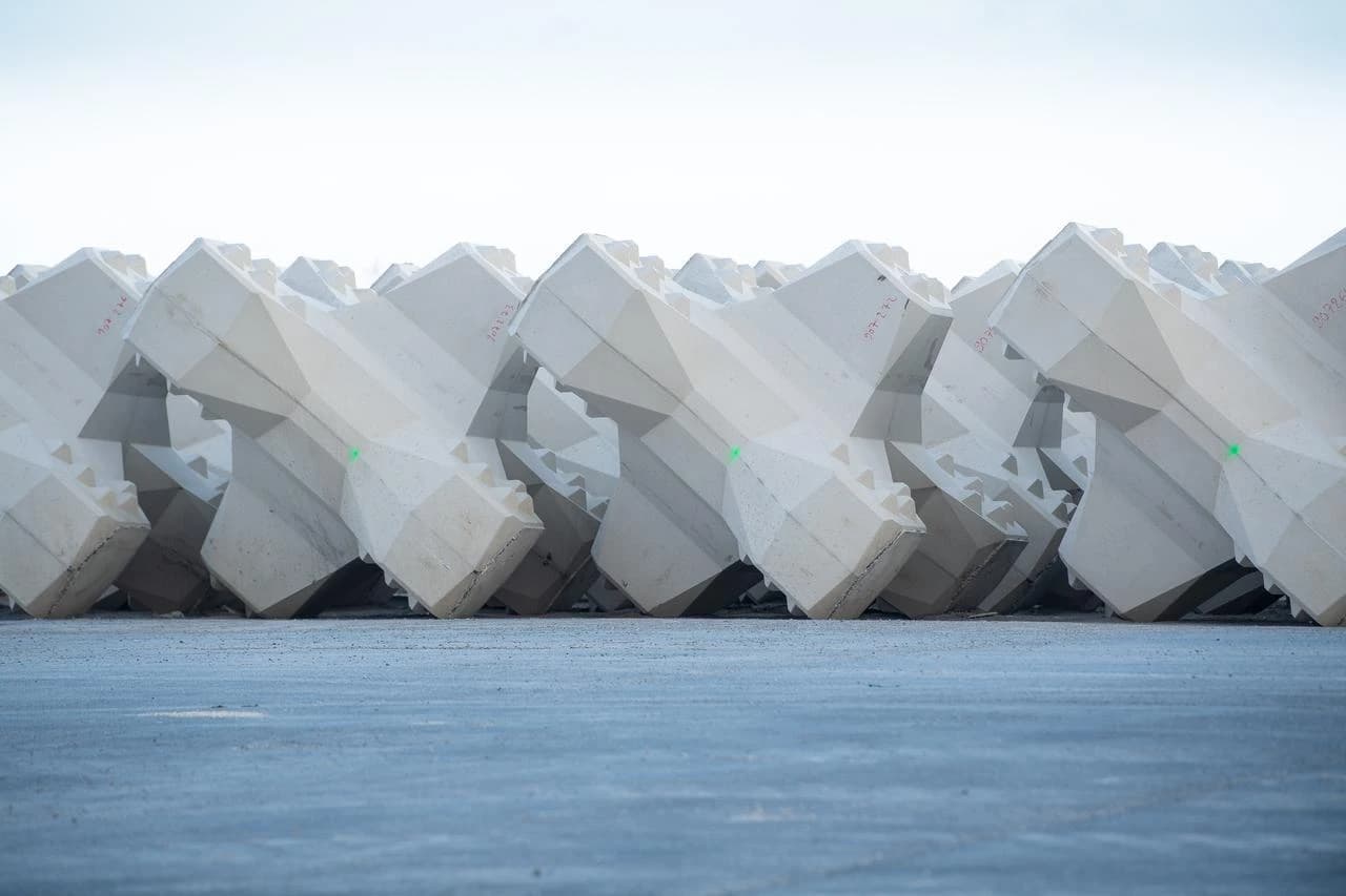 Une longue rangée de blocs de béton gris clair, conçus pour les digues, est alignée sur un sol sombre sous un ciel lumineux. Chaque bloc présente des formes géométriques complexes et de légères marques rouges ou vertes.