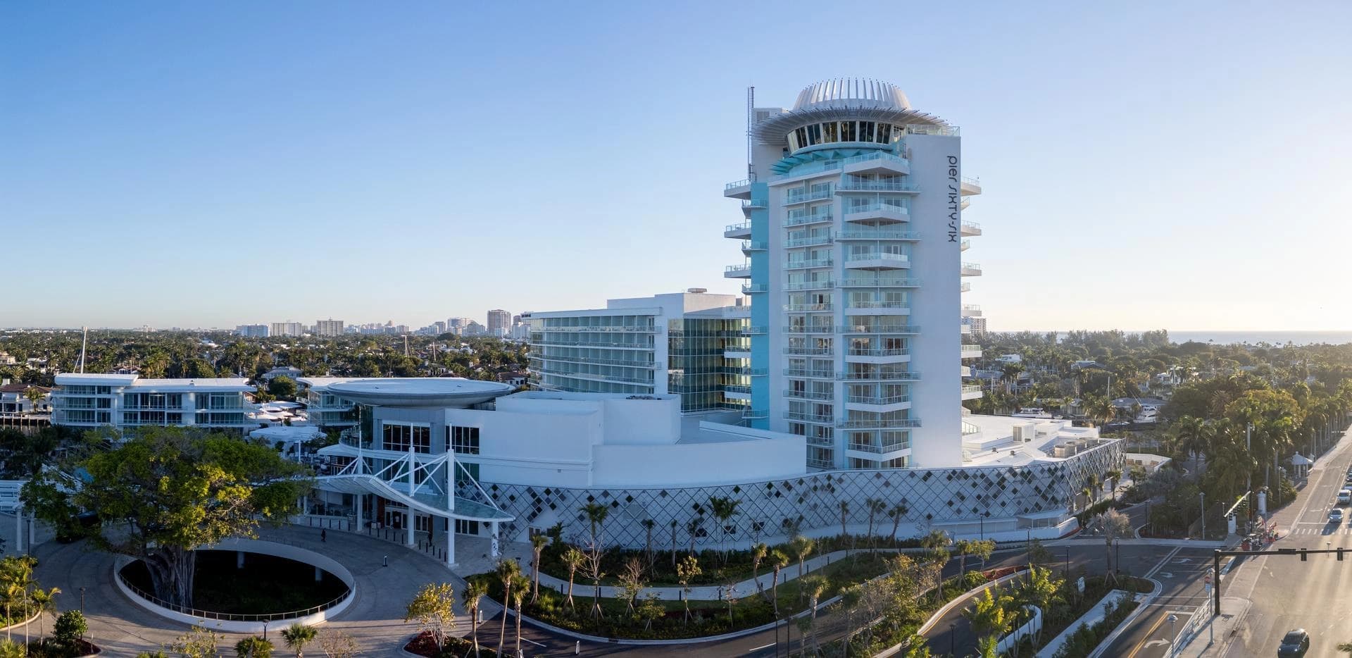 Vue aérienne du moderne hôtel "pier SIXTY-SIX" en blanc et bleu clair, avec une base ornée de motifs géométriques, des balcons en verre et un dôme circulaire au sommet, entouré de palmiers et d'un paysage urbain menant à l'océan lointain sous un ciel dégagé.