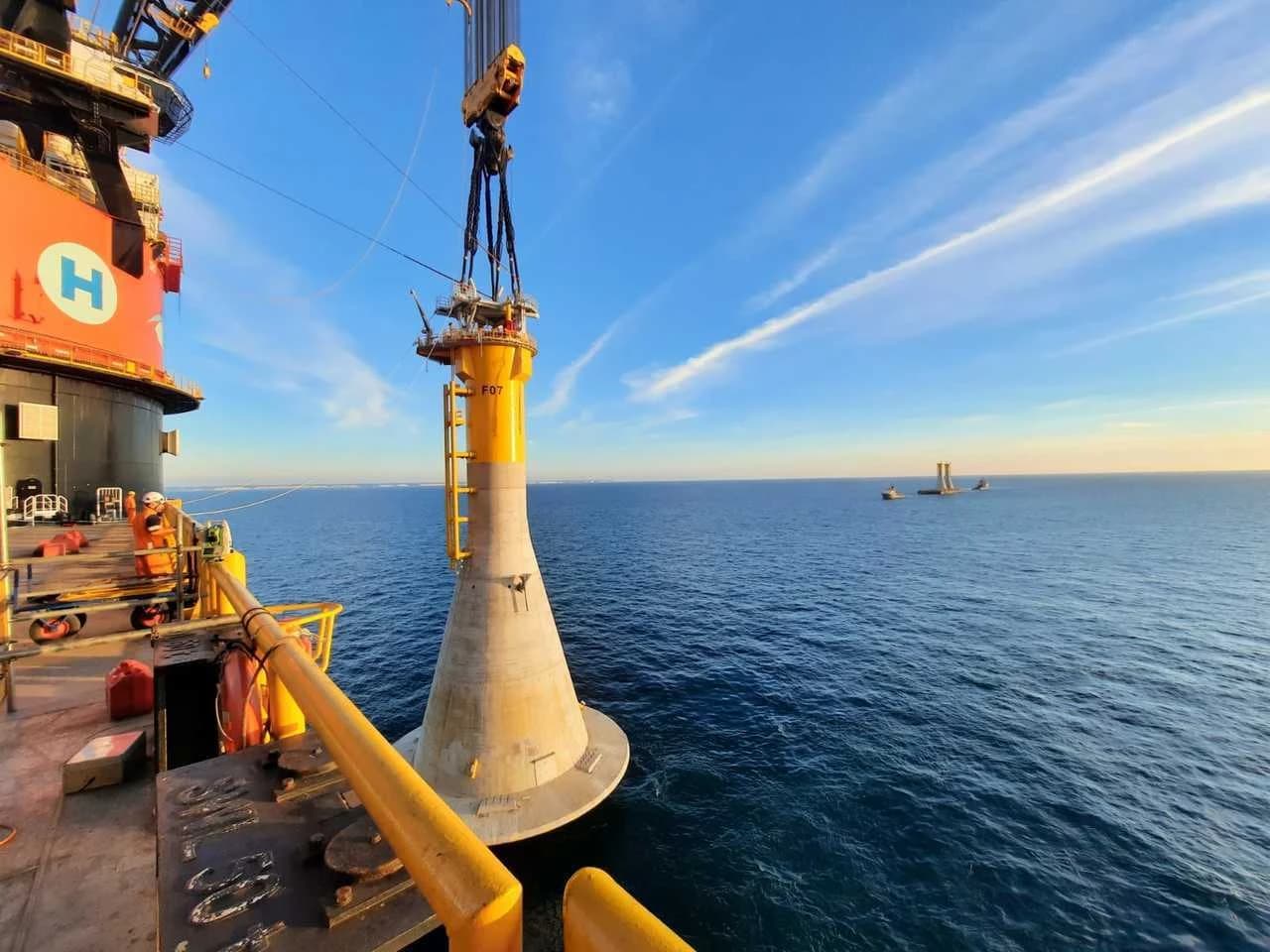Un grand bateau grue orange positionne une massive fondation de turbine éolienne conique jaune et grise, marquée F07, au-dessus de la mer d'un bleu profond sous un ciel clair.