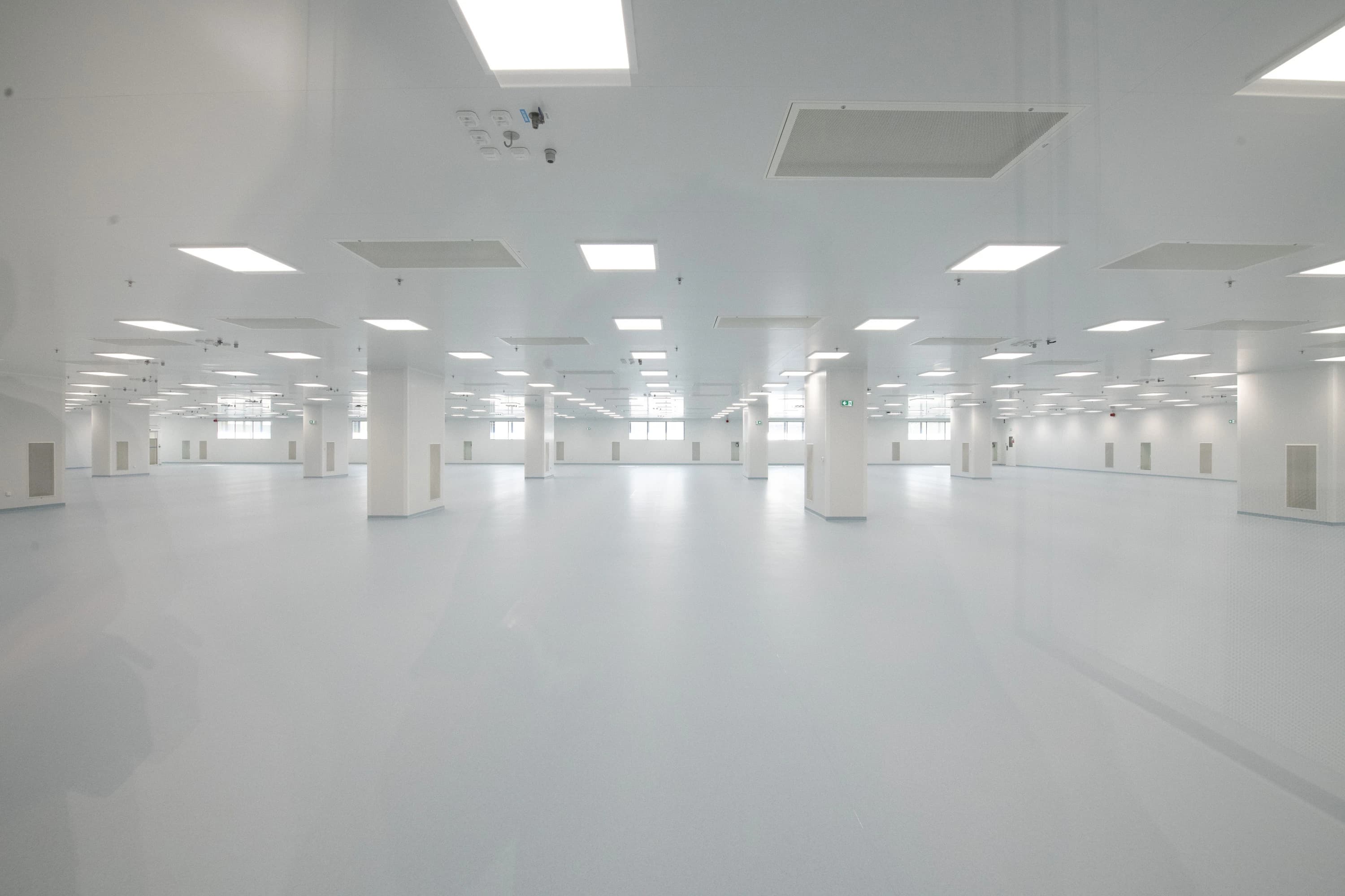 Une vaste salle blanche vide de type salle blanche ou laboratoire, dotée d'un sol gris-bleu clair, de nombreux piliers blancs et de multiples luminaires rectangulaires intégrés au plafond.
