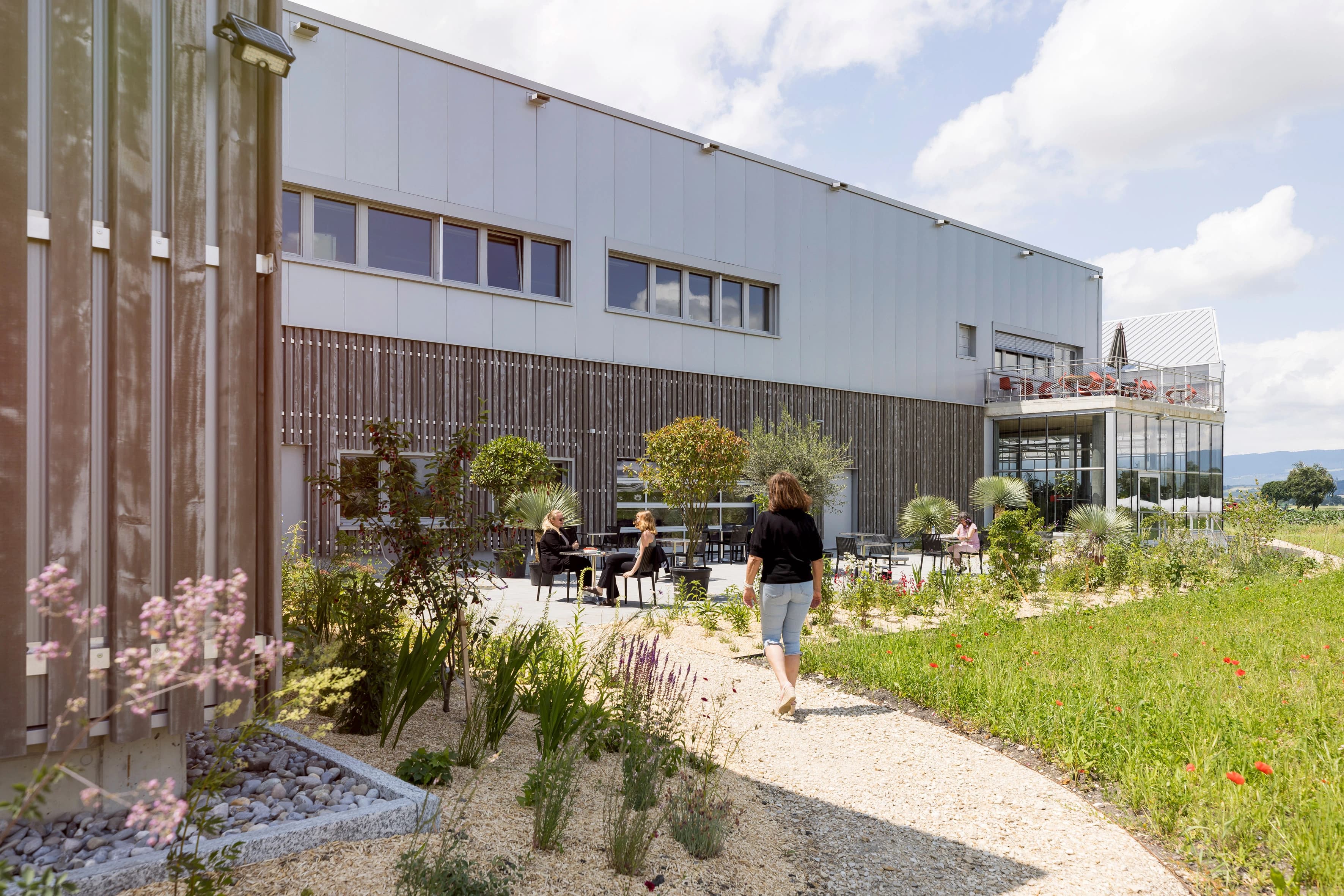 Un bâtiment moderne en panneaux gris et bois sombre avec une terrasse aménagée où des personnes sont assises, et un jardin paysager avec un chemin de gravier menant à un champ vert sous un ciel ensoleillé.