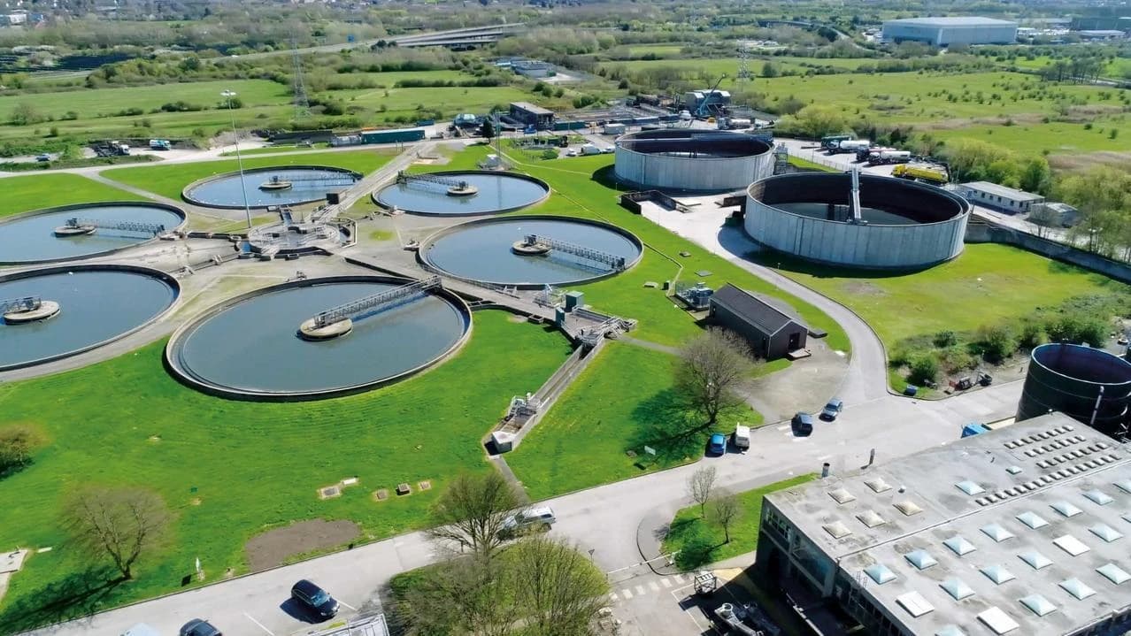 Vue aérienne d'une station d'épuration avec de grands bassins de traitement circulaires et d'autres infrastructures, entourés de pelouses vertes sous un ciel clair.