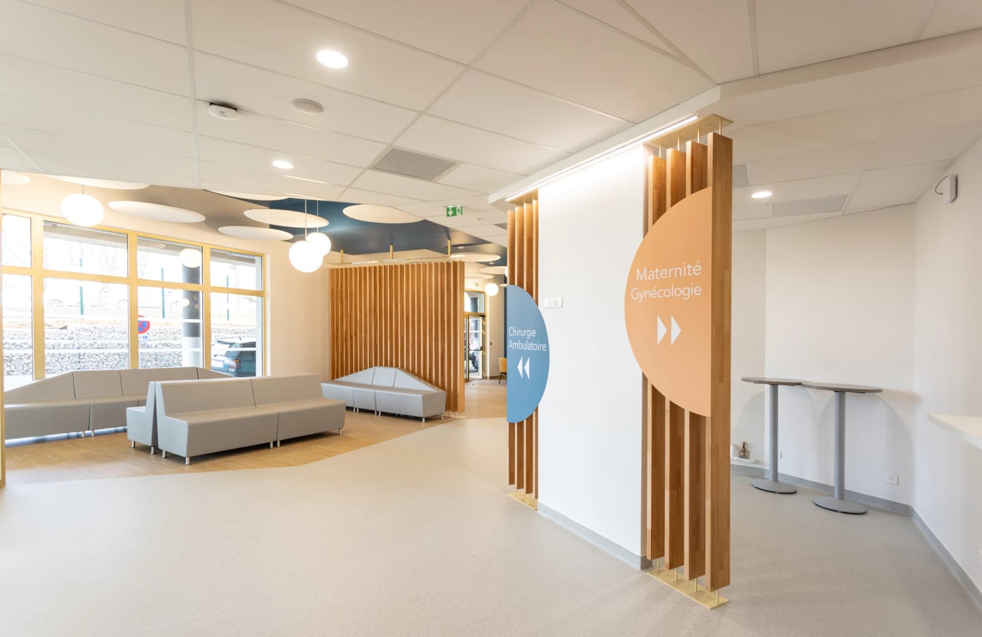 Espace d'attente moderne et lumineux d'un hôpital, avec des canapés gris clair, des cloisons à lattes de bois et de grandes fenêtres. Des panneaux directionnels colorés indiquent la "Maternité Gynécologie" (orange) et la "Chirurgie Ambulatoire" (bleu).
